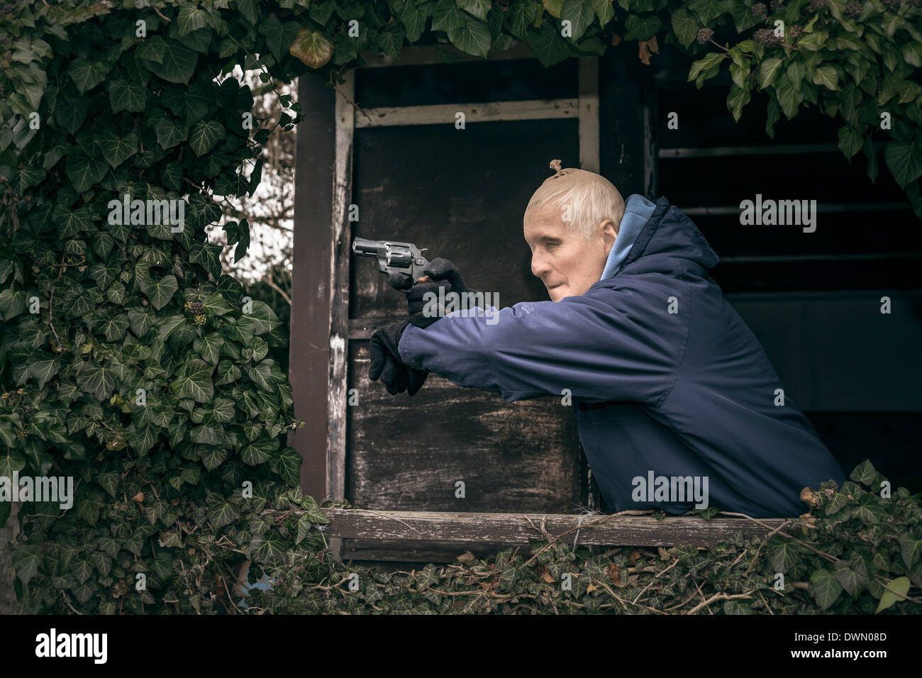 Senior homme masqué évadé visant une arme à feu, se cachant sur de vieux porche envahi par la cabine. Banque D'Images