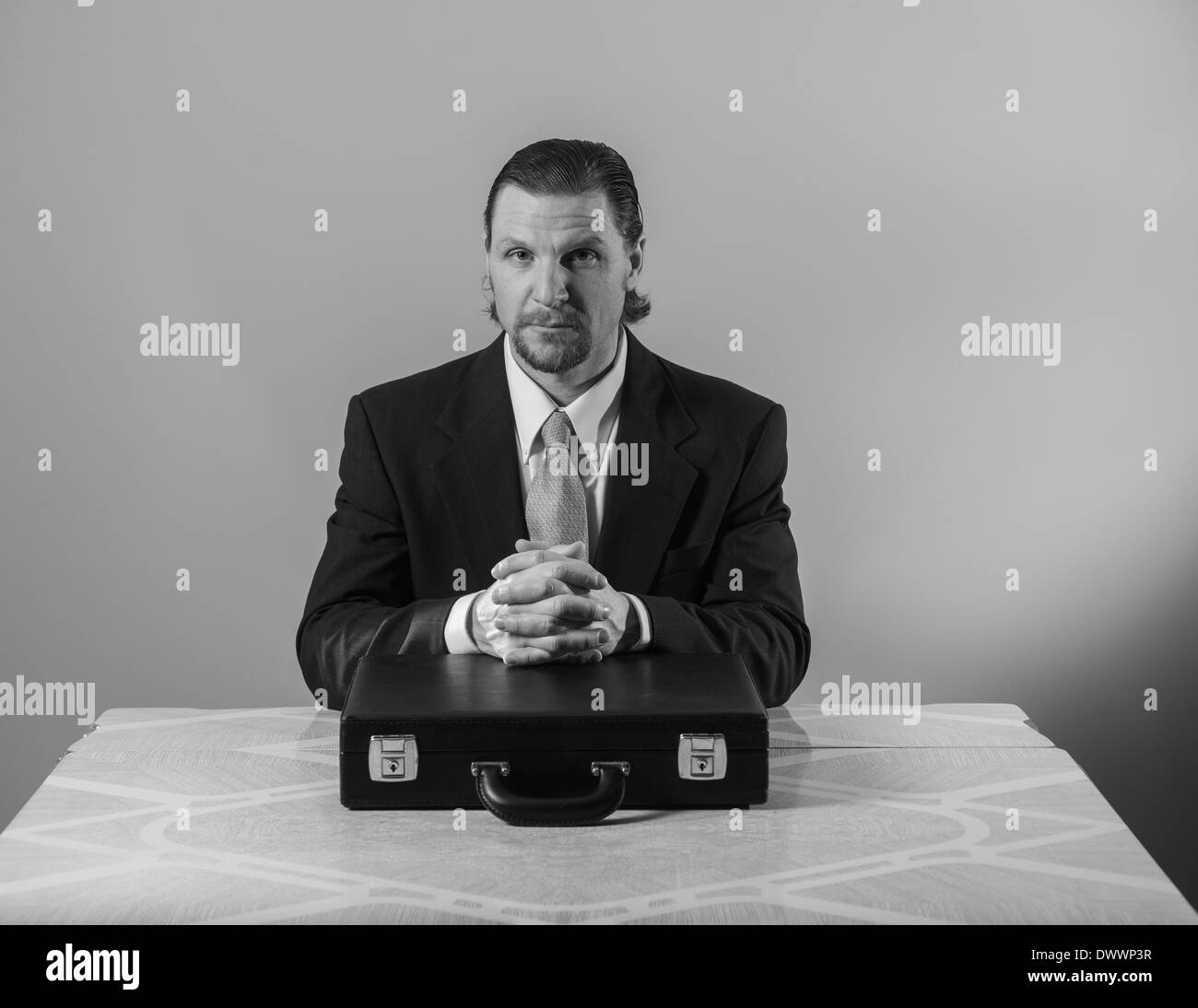 Un 42-year-old woman business man avec une barbiche portant un costume et une cravate est assis à une table de cuisine, les mains croisées sur un attaché-case. Banque D'Images