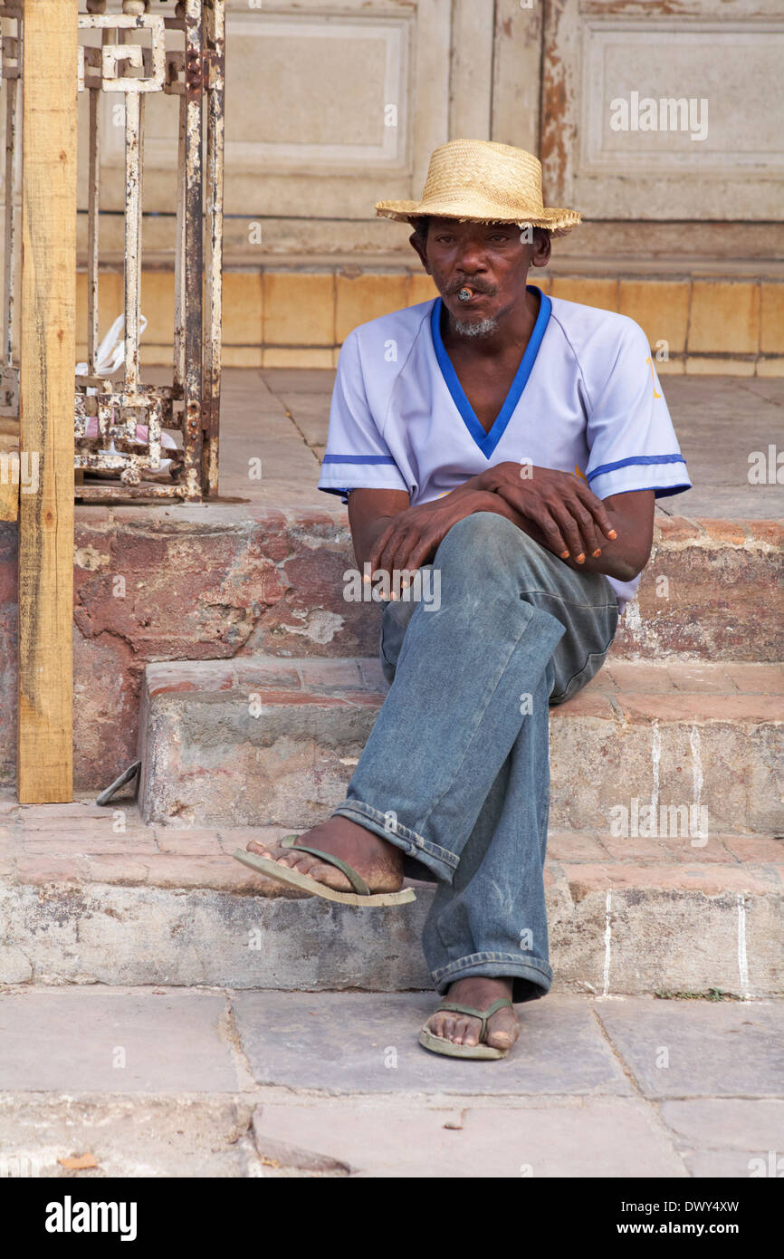 La vie quotidienne à Cuba - afro-caraïbes homme assis sur les marches fumer un cigare à Trinidad, Cuba Banque D'Images