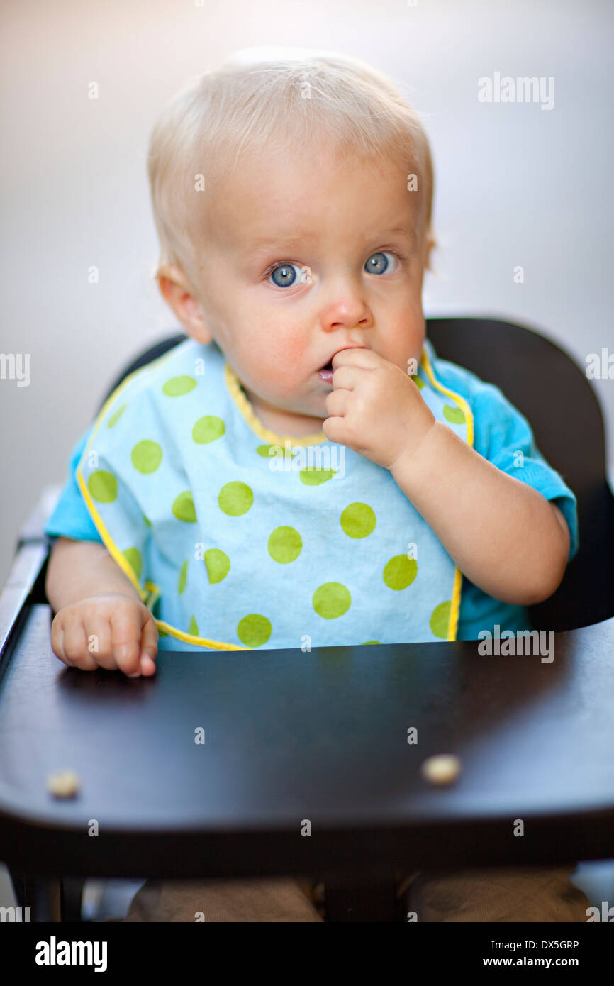 Les yeux écarquillés, baby boy eating cereal in chaise haute avec polka-dot bib, cheveux blonds, yeux bleus, portrait Banque D'Images