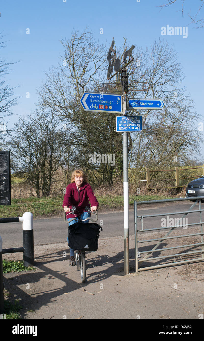 Cycliste femme plus âgée passe un signe sur la National Cycle Route 65 entre York et Beningbrough England UK Mars 2014 Banque D'Images