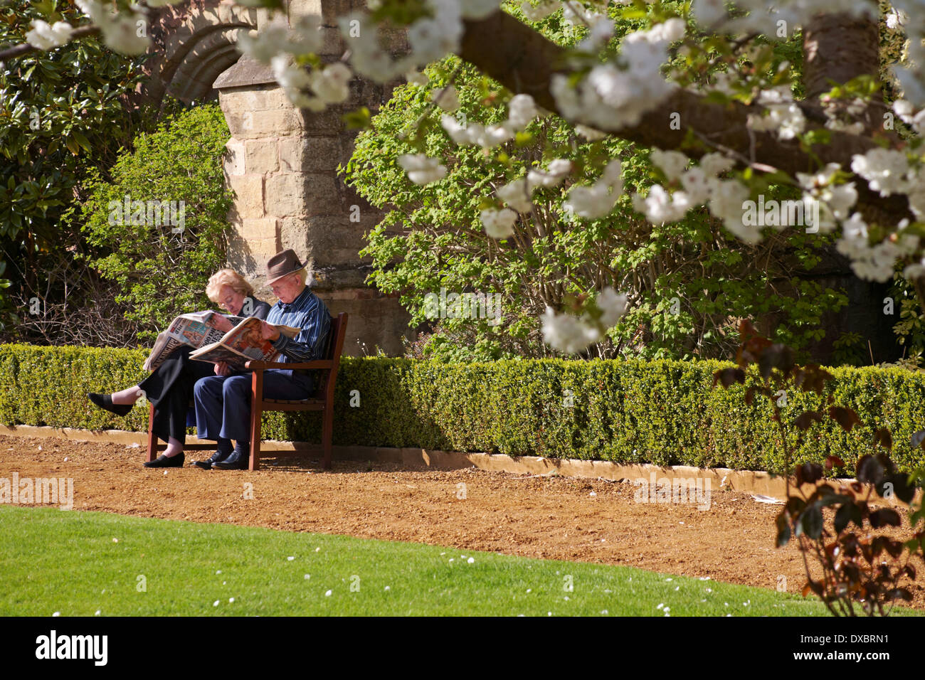 Un couple de personnes âgées lisant des journaux s'est assis sur un banc de fleurs à l'extérieur du prieuré à Great Malvern, Worcestershire, Royaume-Uni, en avril Banque D'Images