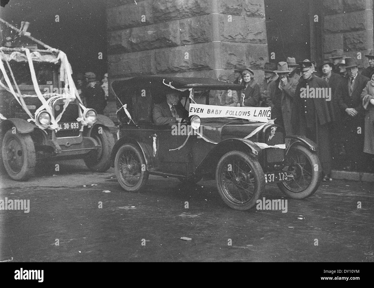 Cette image montre une voiture de bébé Austin (Austin 7), accompagnée d'une affiche qui dit "même bébé suit Lang." Prise en 1934 par Sam Hood, la photographie capture le charme des débuts de la culture automobile en Australie. Banque D'Images