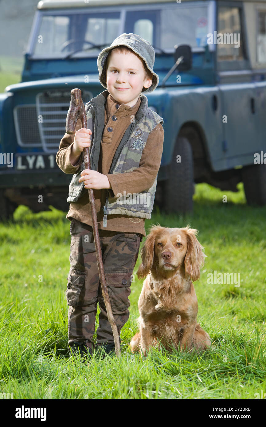 Un garçon de 5 ans dans la campagne typique de l'habillement, à l'extérieur tenant un bâton, avec un chien cocker doré Banque D'Images