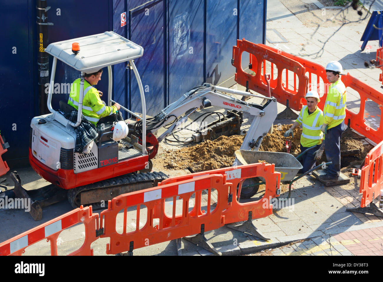 Vue aérienne équipe de trois hommes portant un casque et une veste haute visibilité utilisant une mini pelle creuse creusant le trottoir derrière les barrières rouges Londres Royaume-Uni Banque D'Images