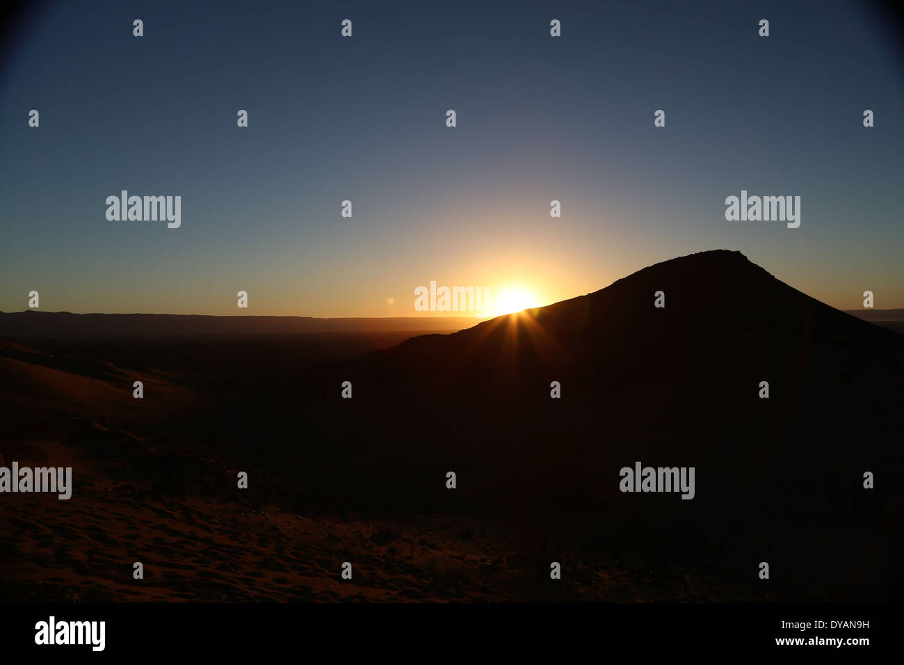 Le lever du soleil sur le baron paysage désertique au Maroc au-delà de l'Atlas des chaînes de montagnes, avec des rayons de soleil une dune de sable qui se chevauchent Banque D'Images