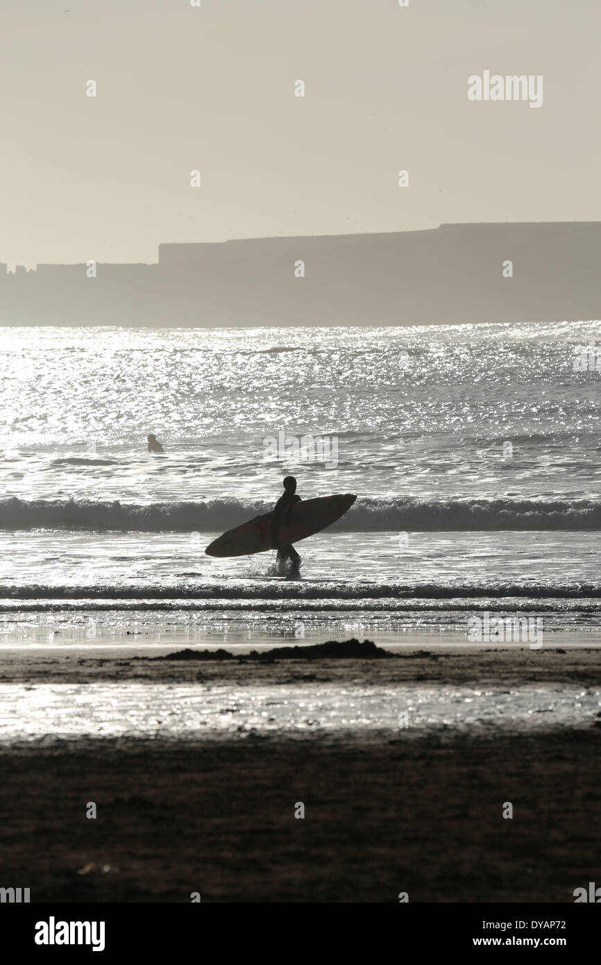 Surfer carrying sa planche de surf le long de la le long du clapotis des vagues d'une plage à Essaouira, Maroc, une fin de soirée Banque D'Images