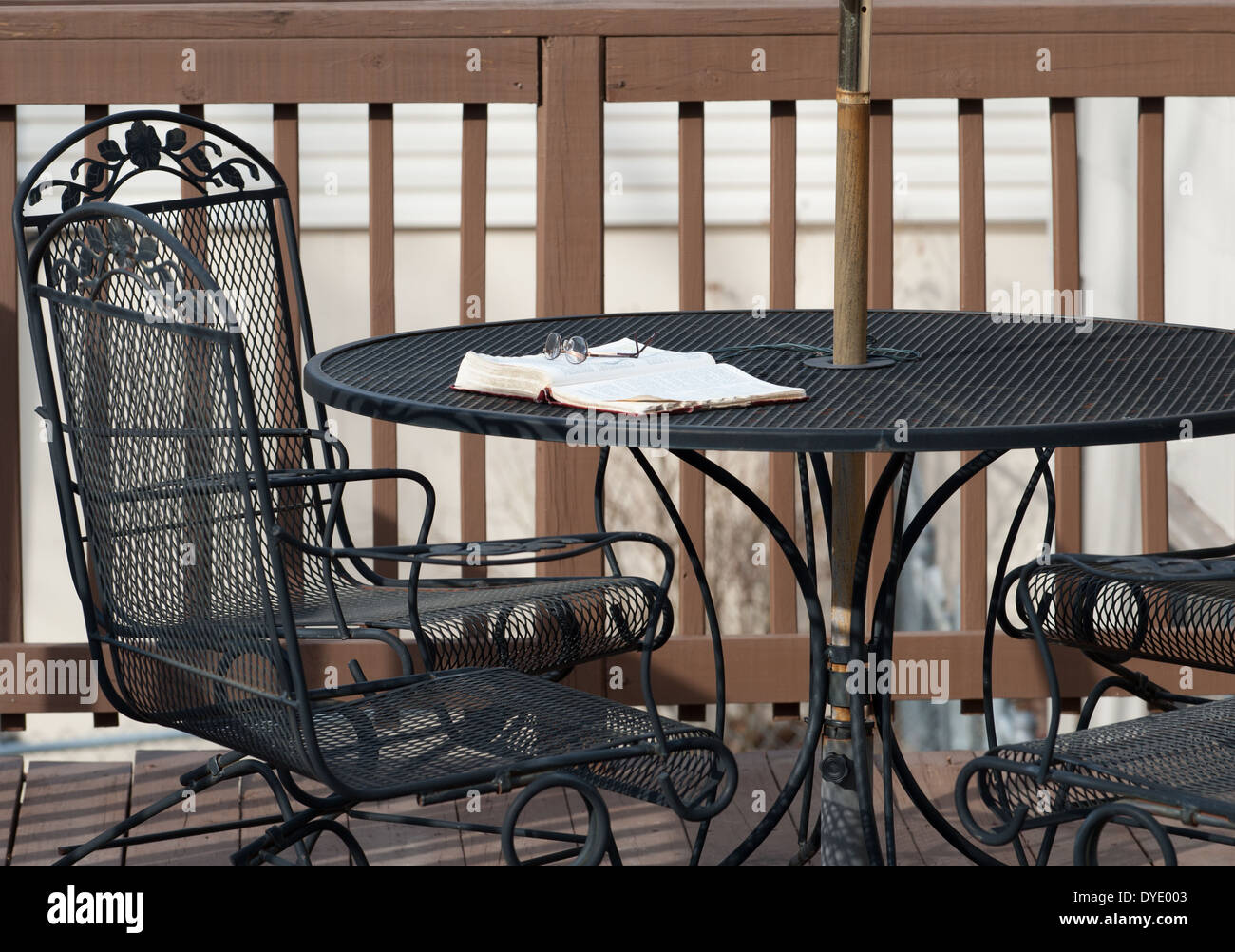 Une paire de lunettes de lecture se trouve au sommet d'une Bible ouverte portant sur une table en fer forgé sur une terrasse en bois avec chaises assorties. Banque D'Images
