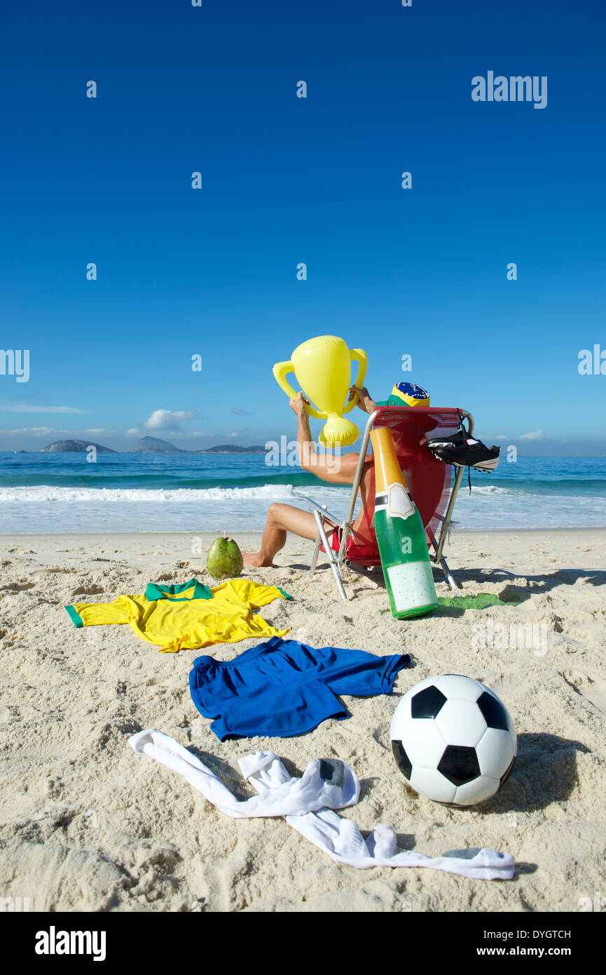 Joueur de football brésilien, Champion de célébrer en vous relaxant dans Rio de Janeiro chaise de plage avec trophée, ballon de soccer, champagne Banque D'Images