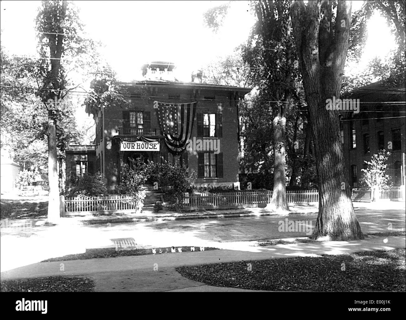 Cette photographie de Bion Whitehouse montre une maison à Keene, dans le New Hampshire, ornée d'une banderole patriotique. L'image capture la cour avant avec une clôture et des ormes, mettant en valeur un cadre festif et historique dans la ville de Keene. Banque D'Images