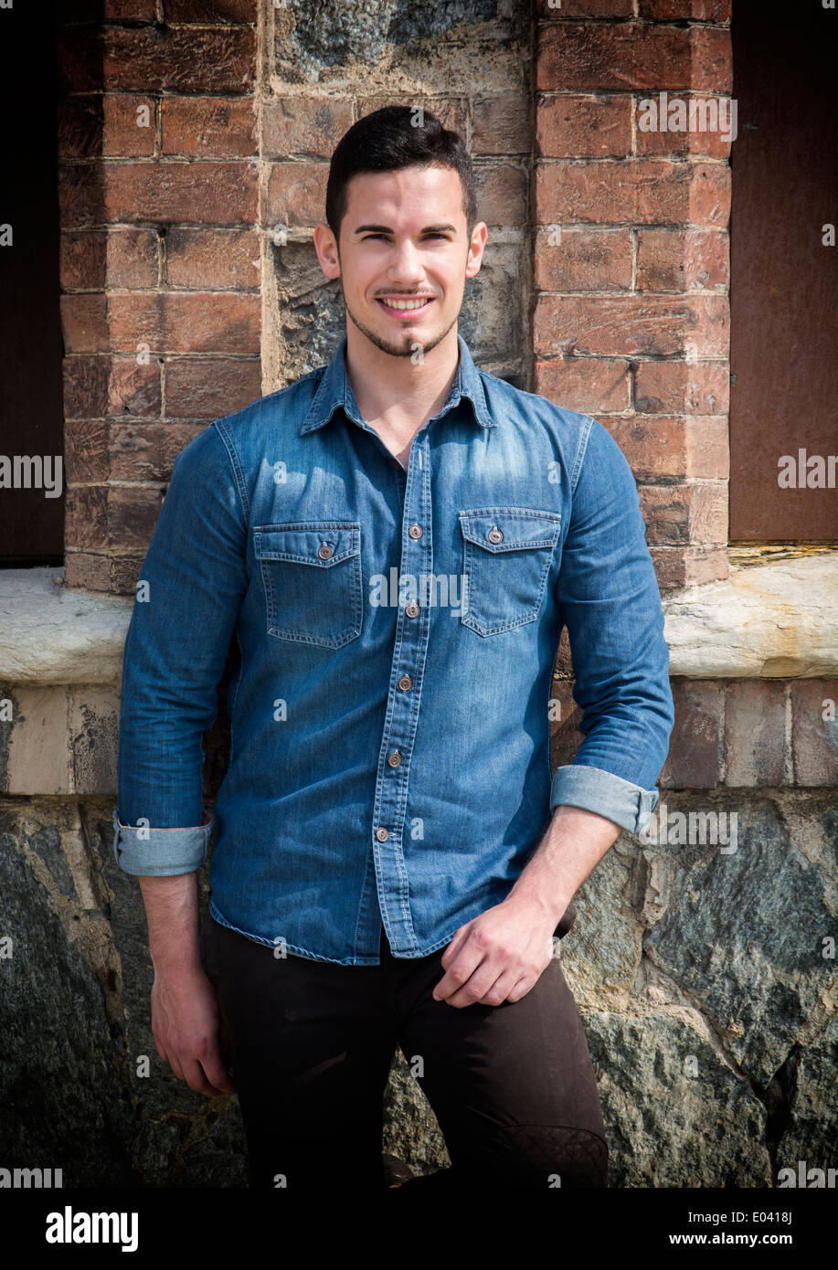 Beau jeune homme avec chemise en jean à l'extérieur debout en face de mur de briques, smiling Banque D'Images