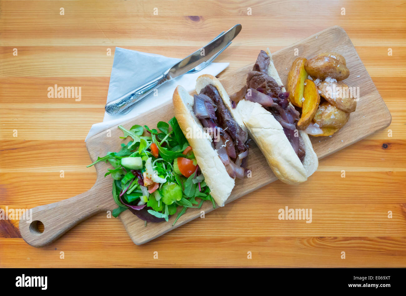 English l'heure du déjeuner collation, un steak et oignon rouge baguette avec de la salade et des quartiers de pommes de terre frits servi sur une planche en bois Banque D'Images