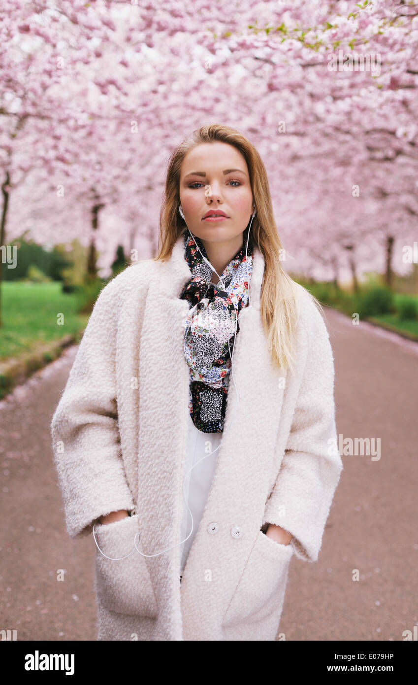 Jolie jeune femme model wearing long manteau d'écouter de la musique tout en fleur de printemps jardin. Banque D'Images