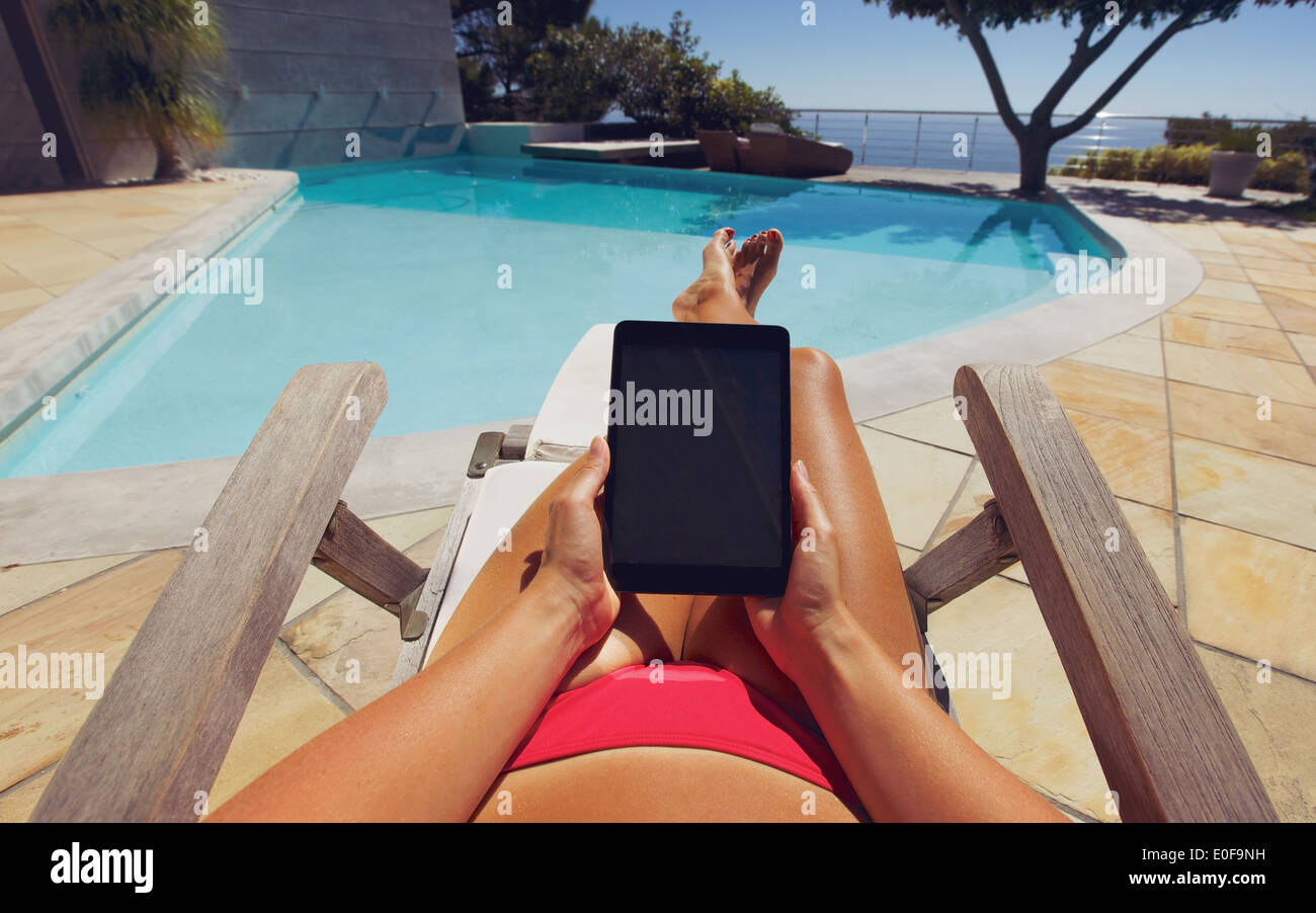 Jeune femme à l'aide d'un tablet PC, près de la piscine. POV de l'utilisateur. Modèle féminin de détente sur un transat holding digital tablet. Banque D'Images