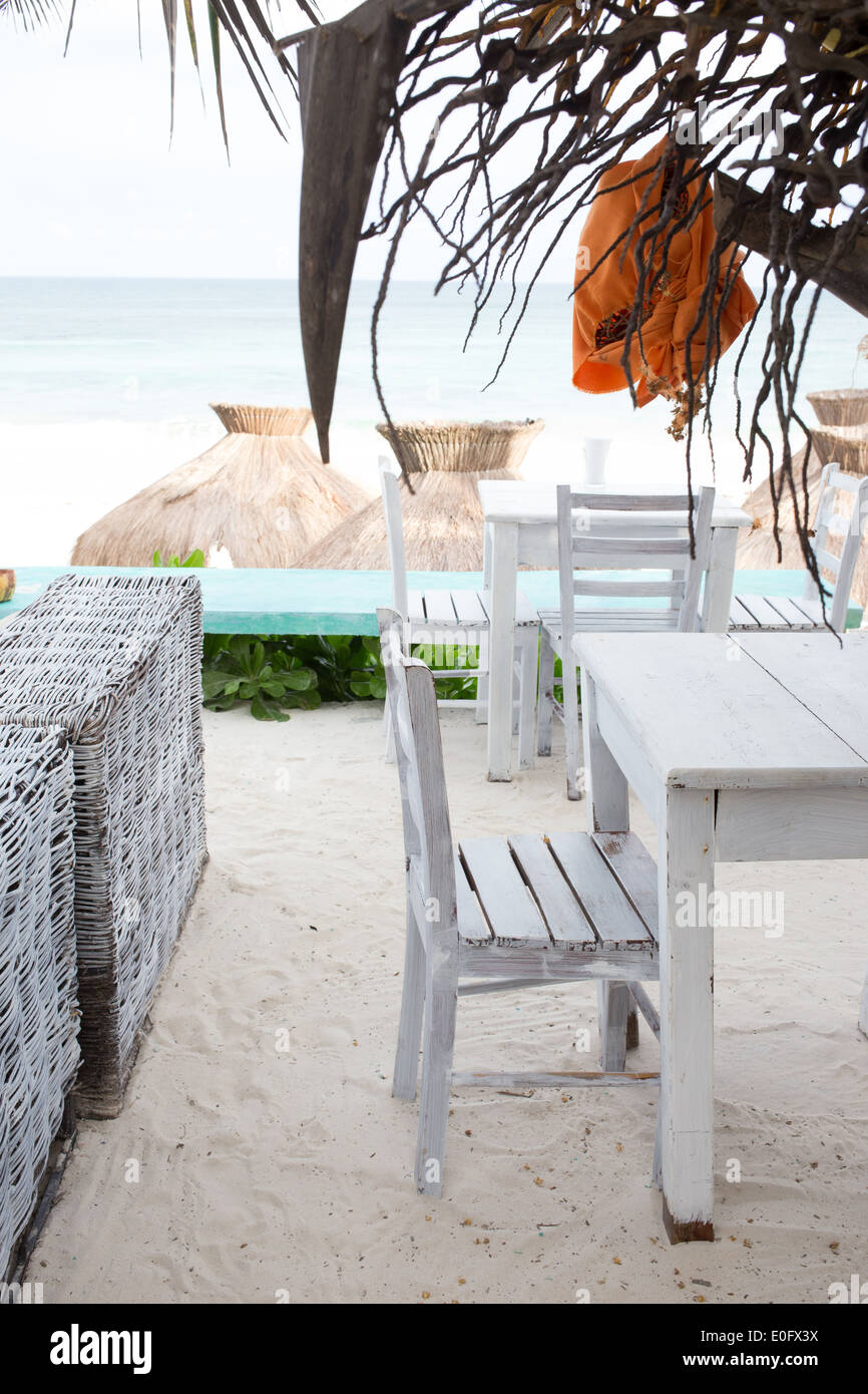 Table et chaises en bois blanc sur la surface sablonneuse avec ocean et toits de paille à l'arrière-plan de Tulum, Mexique Banque D'Images