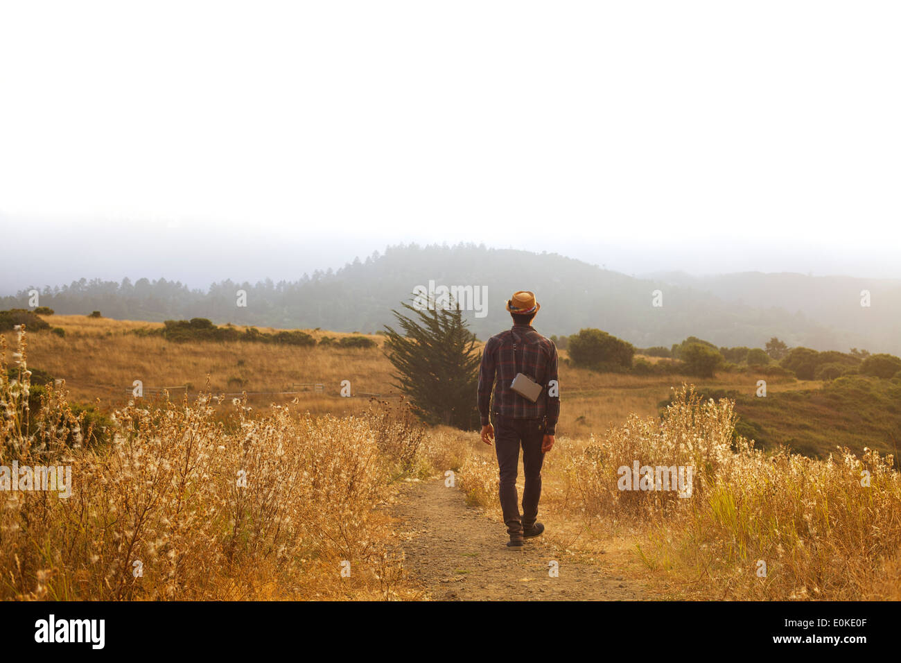 Un homme avec un chapeau de paille et une terre marche le long de la caméra d'un chemin dans un champ d'herbe d'or à sec. Banque D'Images