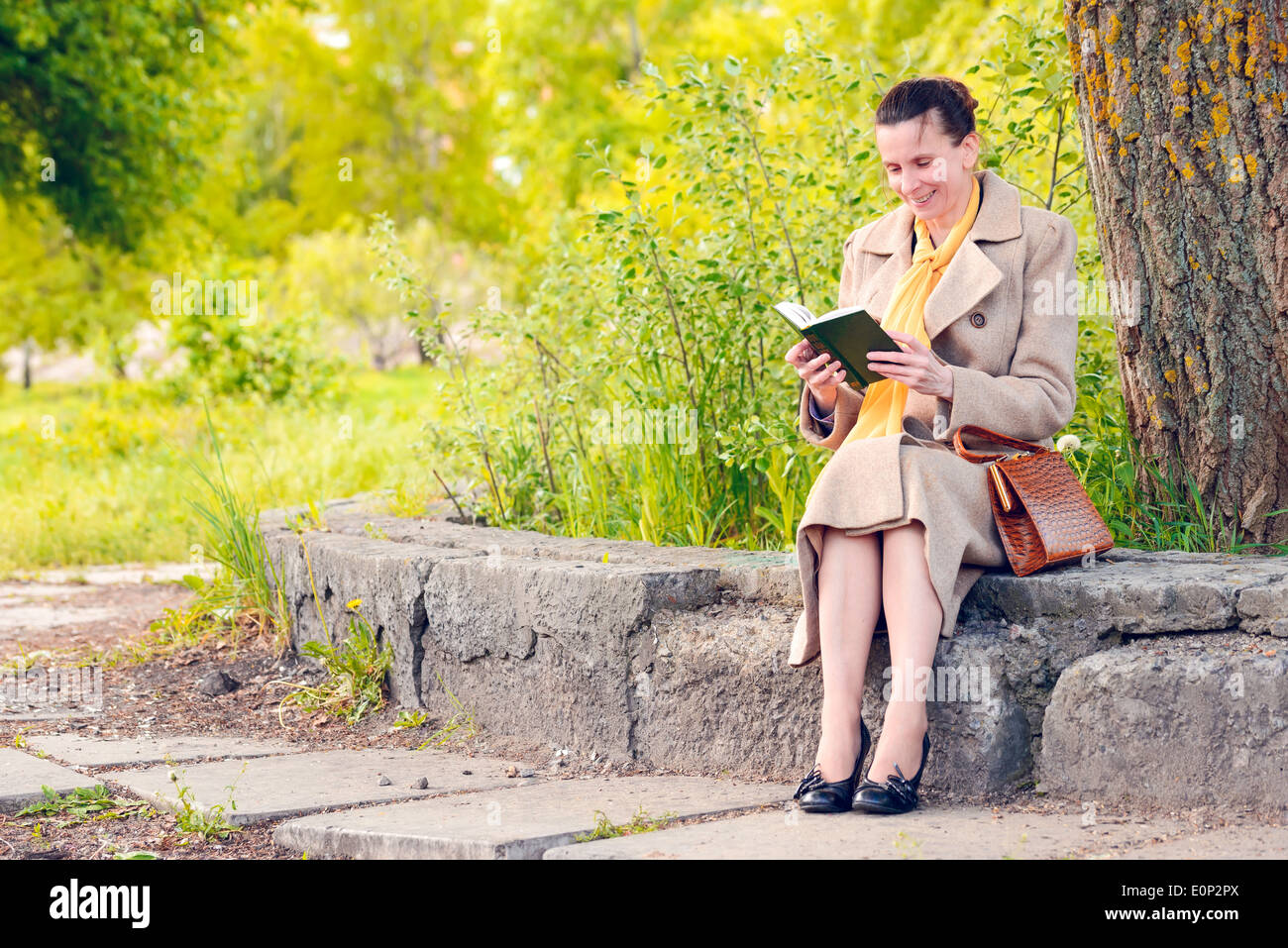 Femme élégante assise sur un mur de pierre au cours d'une journée de printemps ensoleillée, et la lecture d'un livre drôle Banque D'Images
