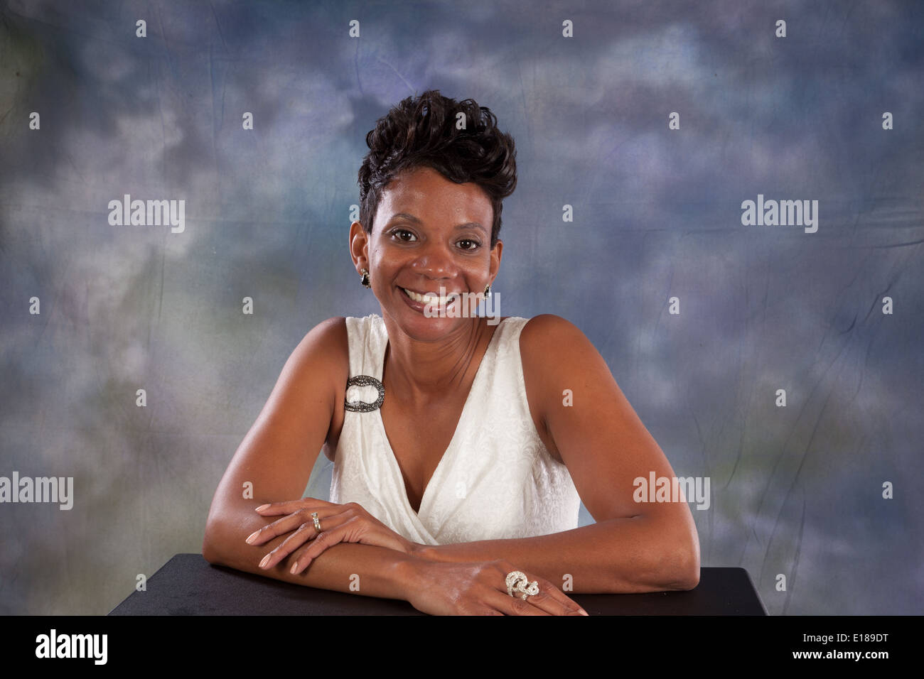 Jolie femme noire en robe blanche, assis avec ses bras sur une table, souriant à la caméra avec plaisir et joie Banque D'Images