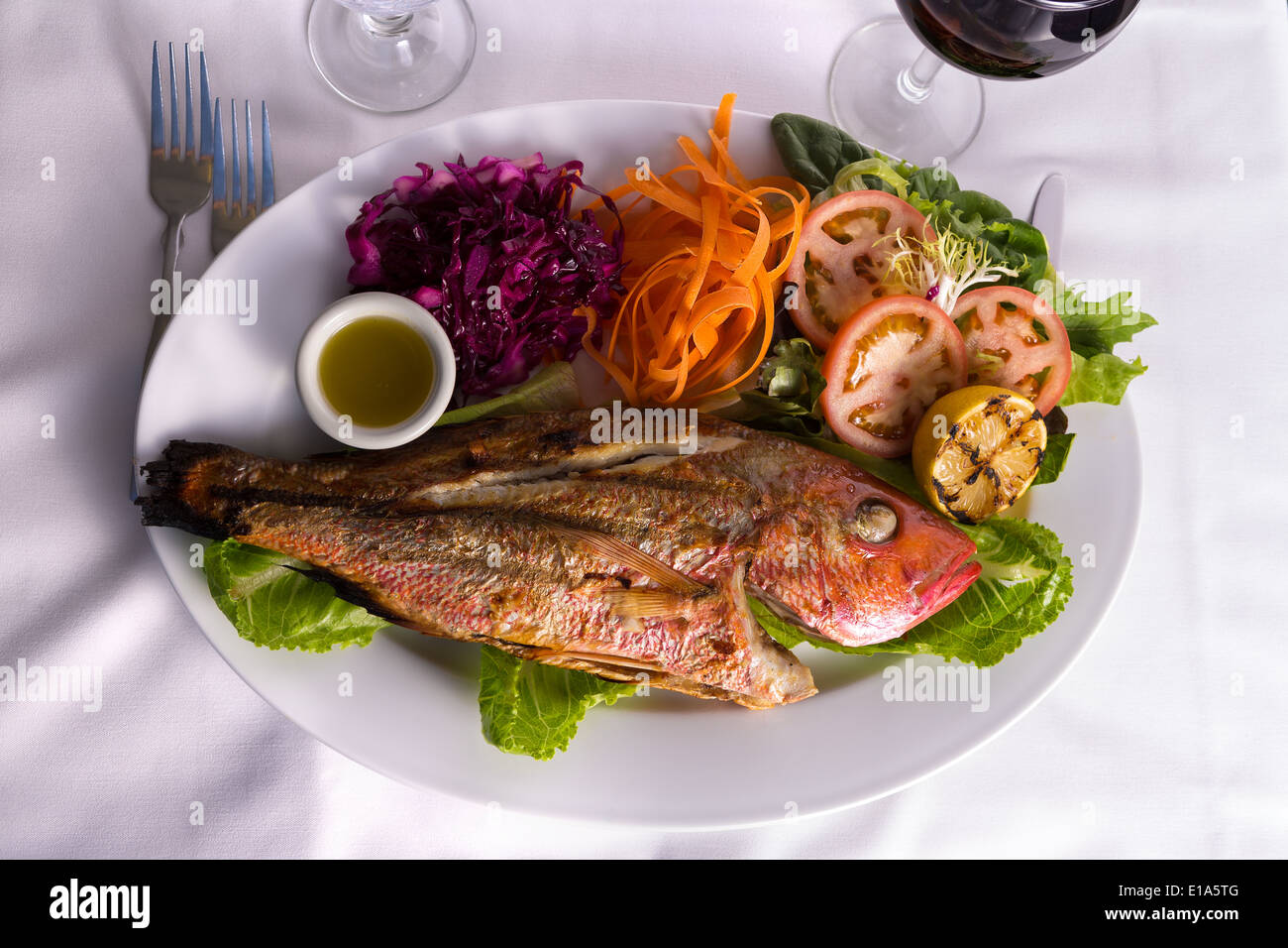 Petit poisson tilapia servi dans une assiette de légumes et sauce de poisson complimenté avec vigne rouge Banque D'Images