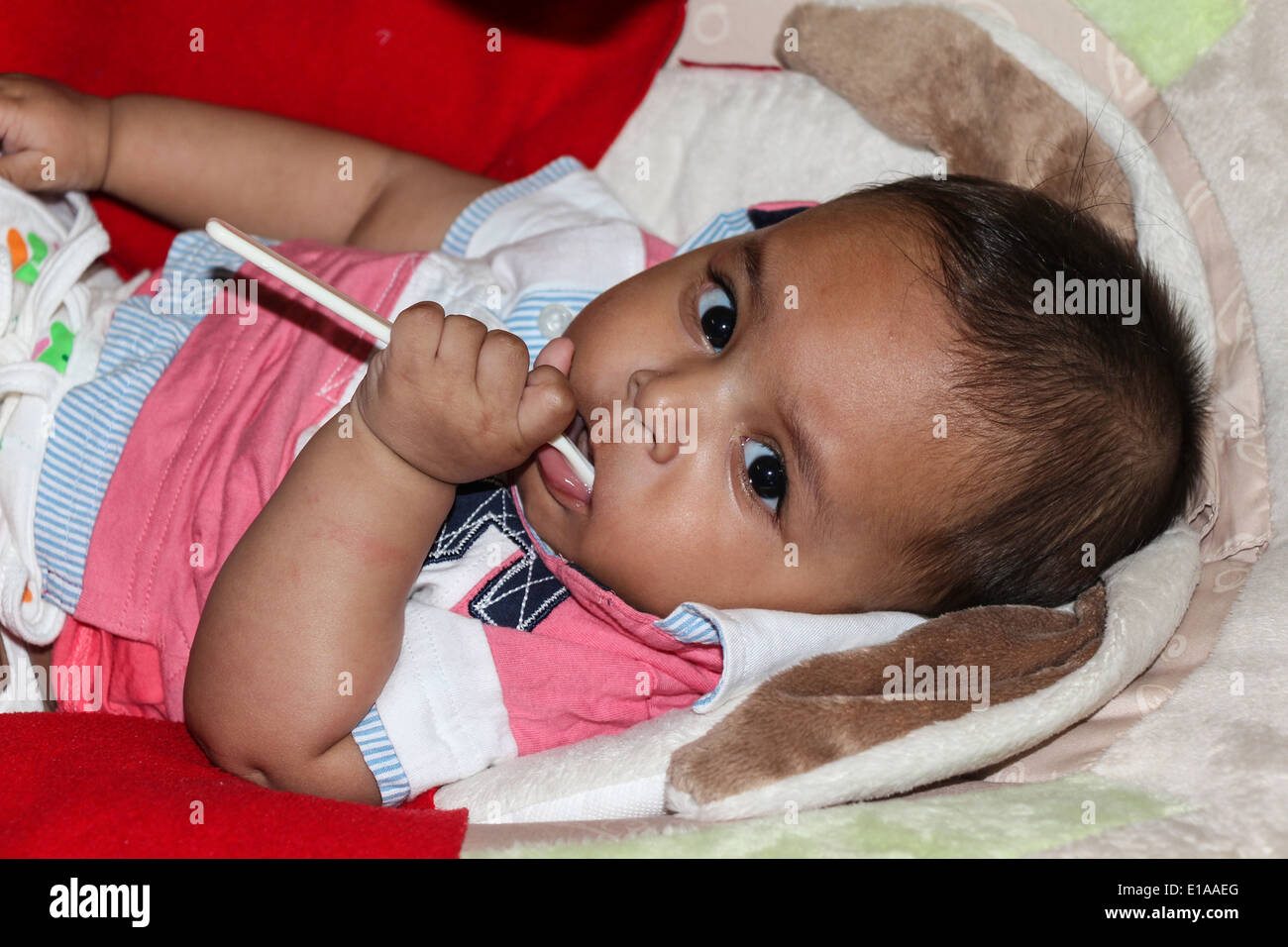 Un petit garçon tenant une cuillère en plastique dans sa bouche tout en étant allongé d'un lit bébé. Le bébé est un petit garçon indien. Banque D'Images