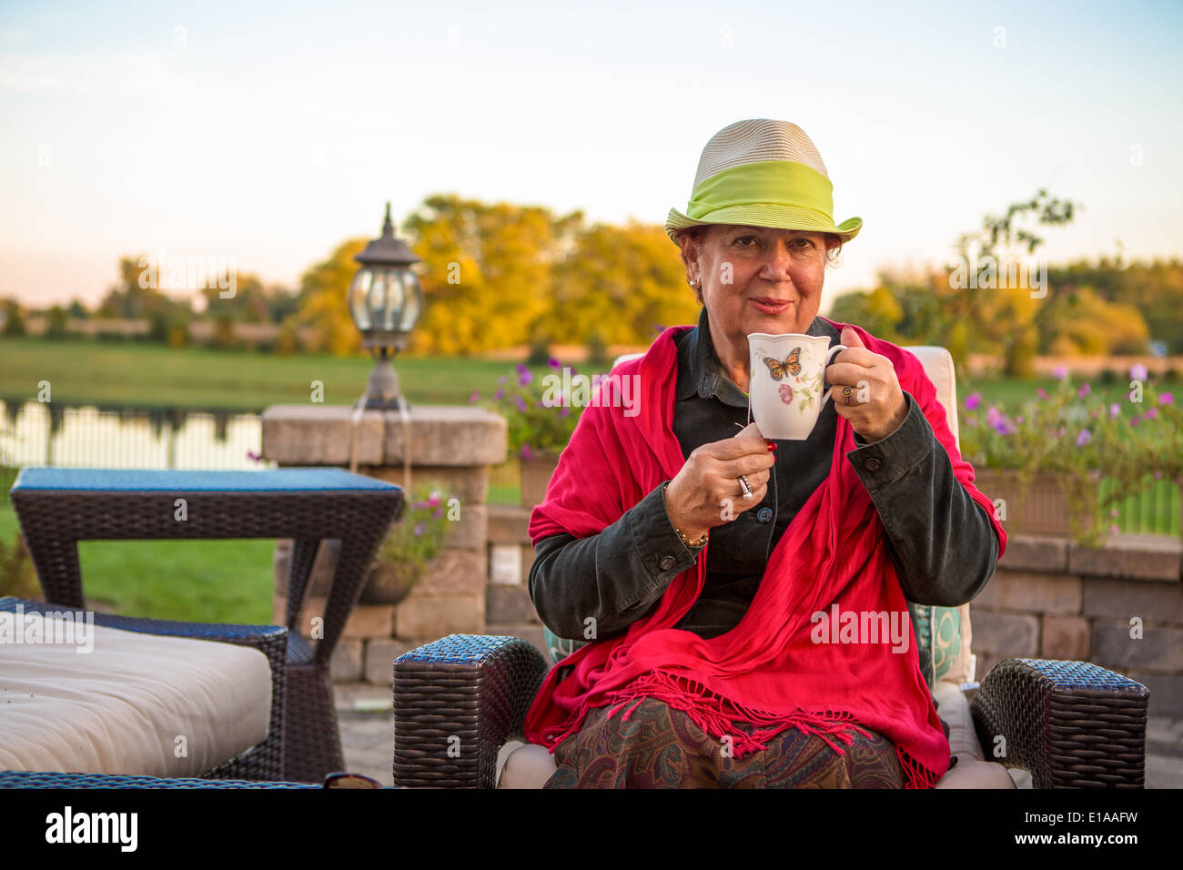 Senior woman avec une paille chapeau vert, assis à la terrasse et en montrant son thé chaud, peut-être qu'elle veut rester au chaud Banque D'Images