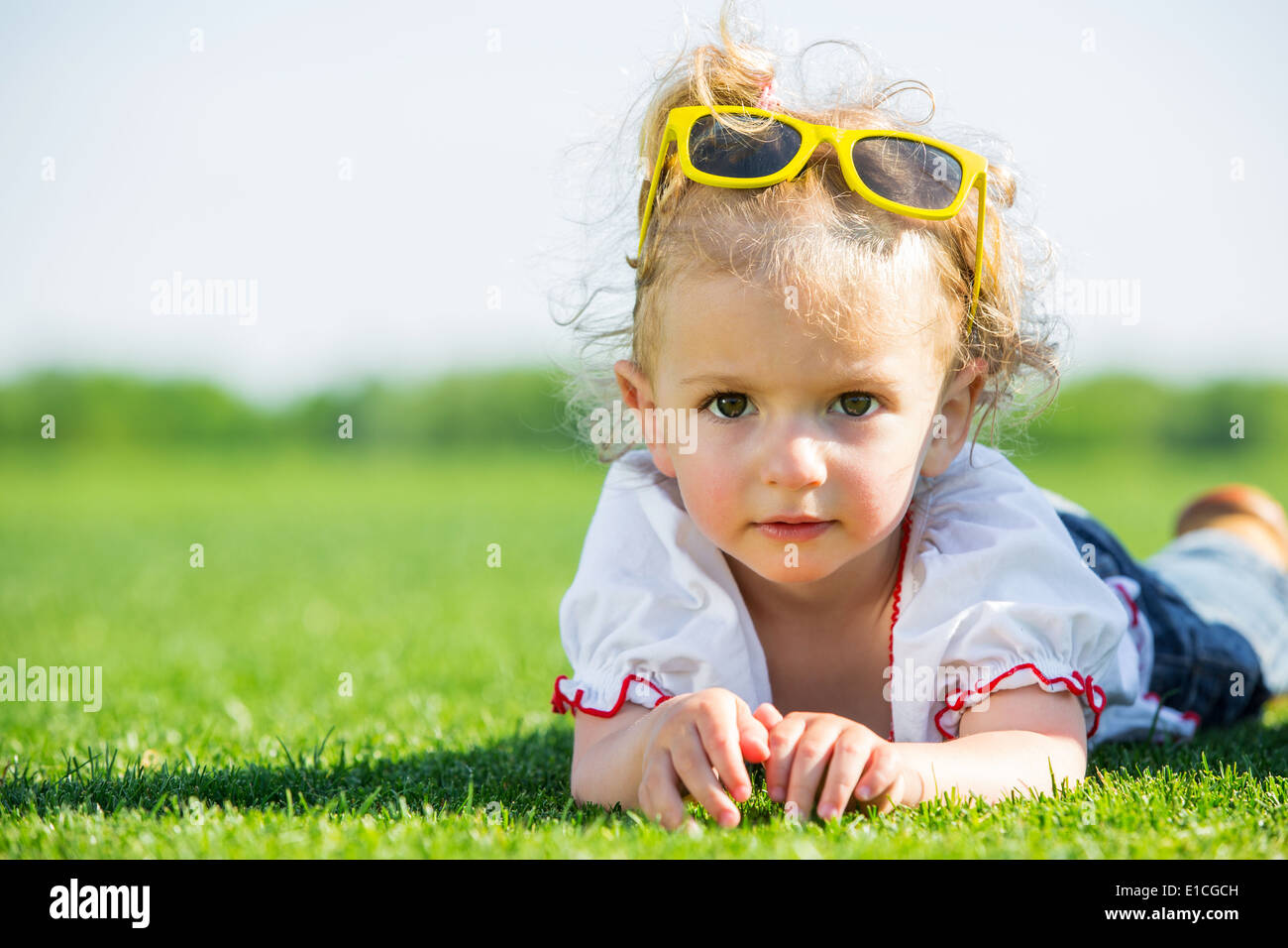Jolie petite fille avec des lunettes de soleil jaune sur le dessus de sa tête, allongé sur une herbe verte fraîche dans un champ Banque D'Images