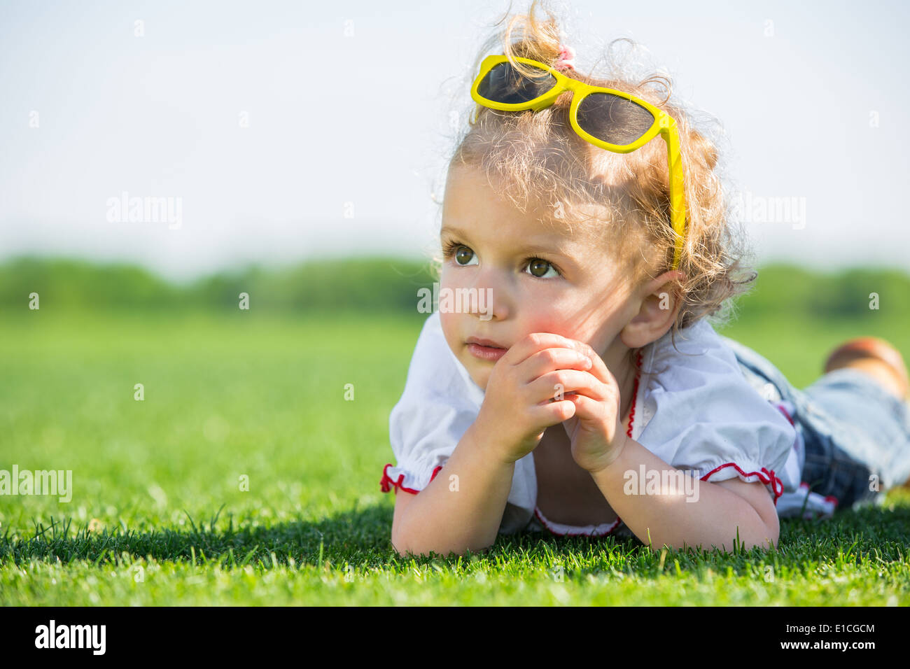 Jolie petite fille avec des lunettes de soleil jaune sur le dessus de sa tête, allongé sur une herbe verte fraîche dans un champ Banque D'Images