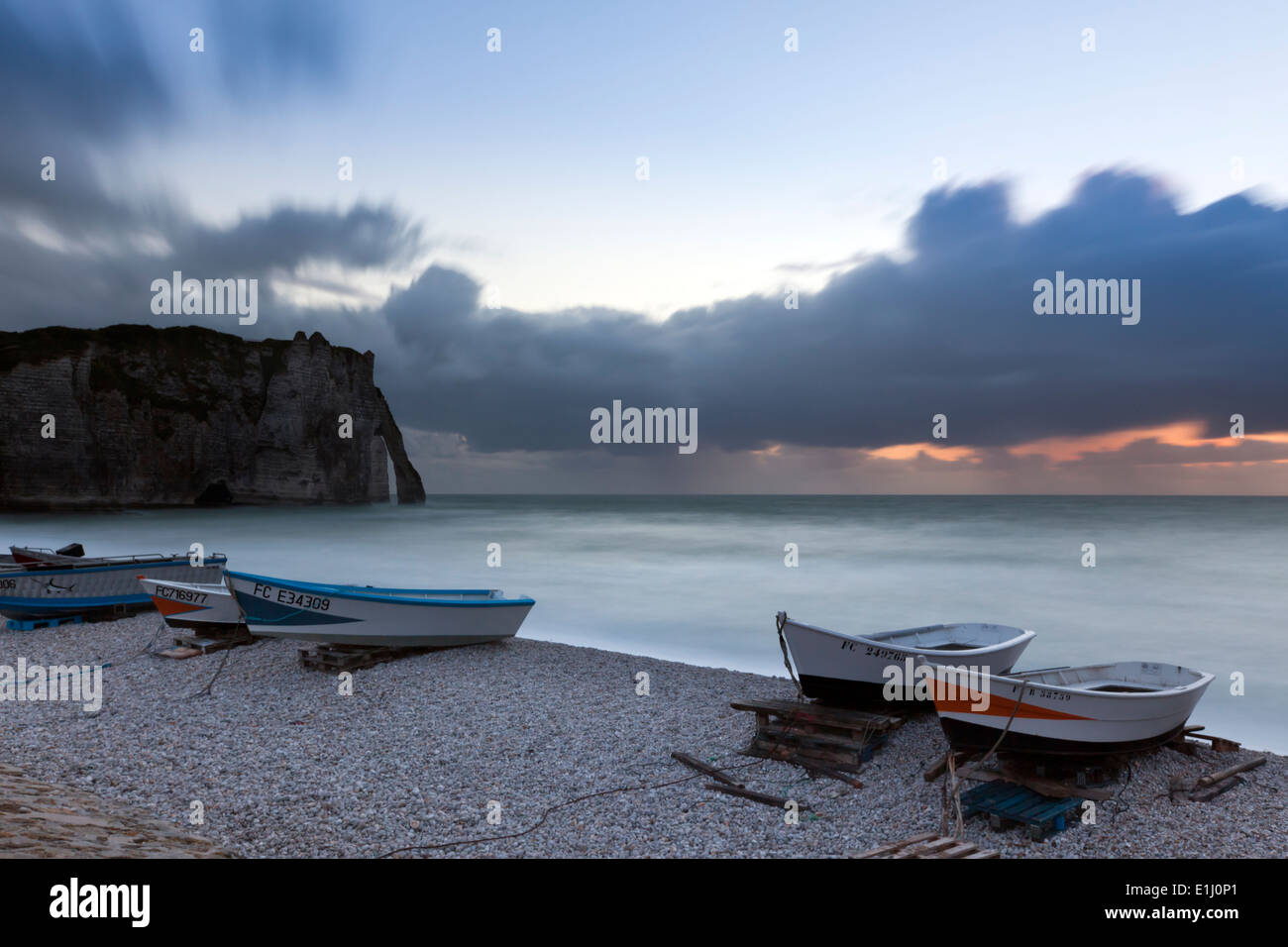 Bateaux sur la plage d'Etretat, Normandie, au coucher du soleil. Porte d'Aval, arche naturelle en arrière-plan. Longue exposition. Banque D'Images