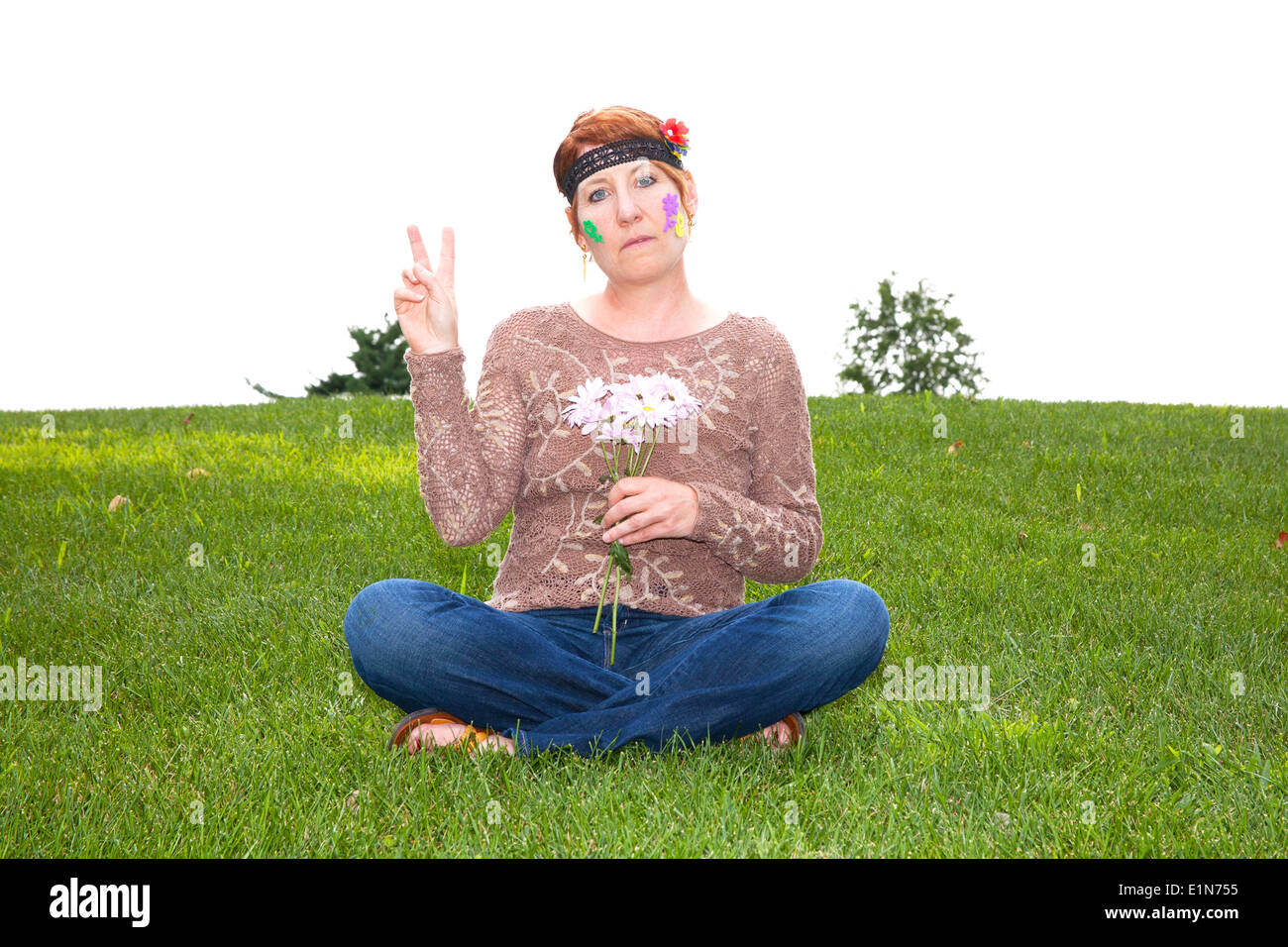 Femme mature habillé en années 60 flower enfant assis dans l'herbe et les pâquerettes maintenant un signe de paix clignotant Banque D'Images