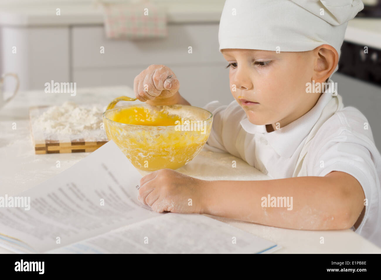 Mignon petit garçon dans un tablier blanc et chefs toque la lecture d'une recette comme il cuisine dans la cuisine le mélange des ingrédients dans un bol Banque D'Images