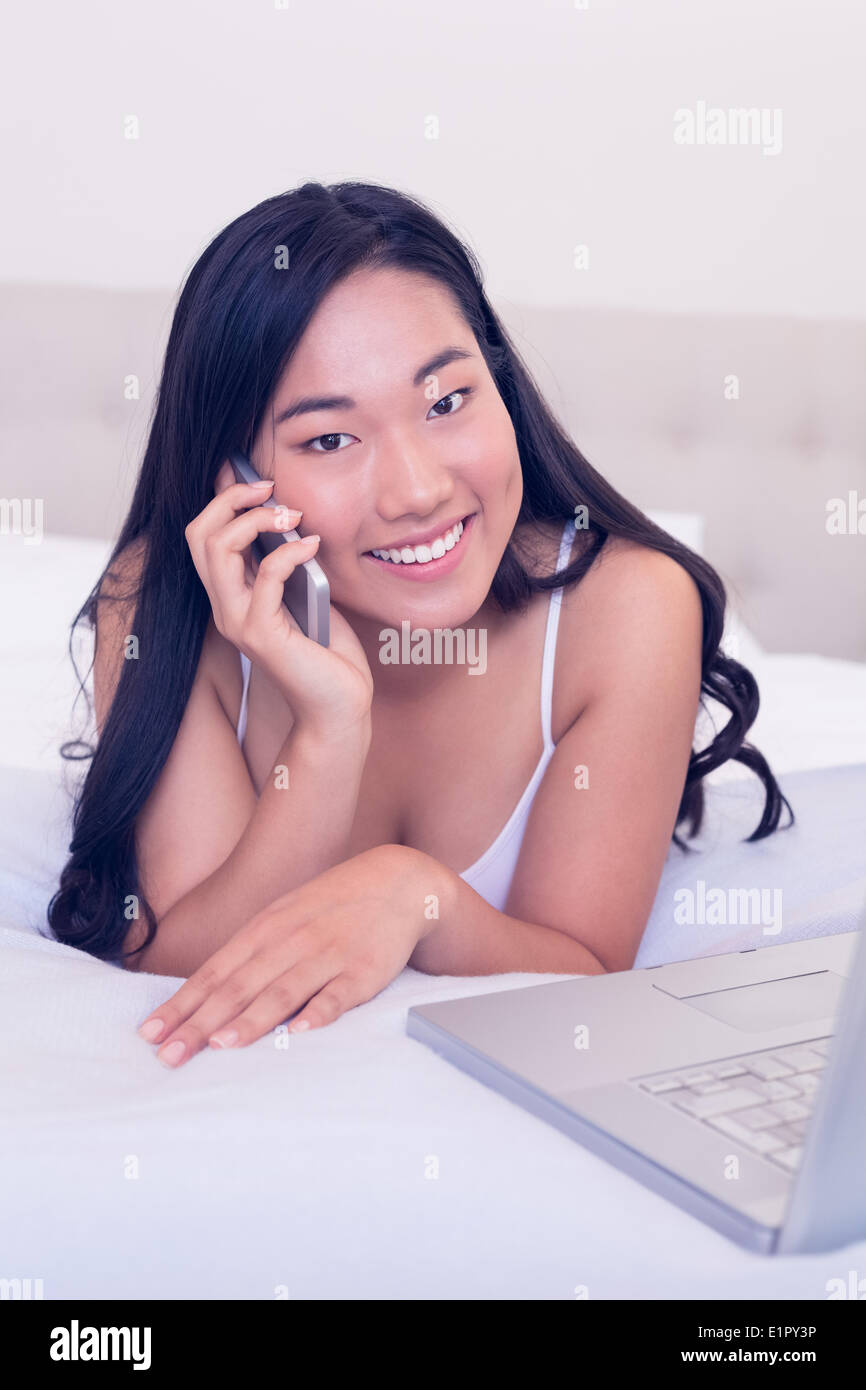 Serveuse asiatique smiling at camera lying on bed talking on phone Banque D'Images