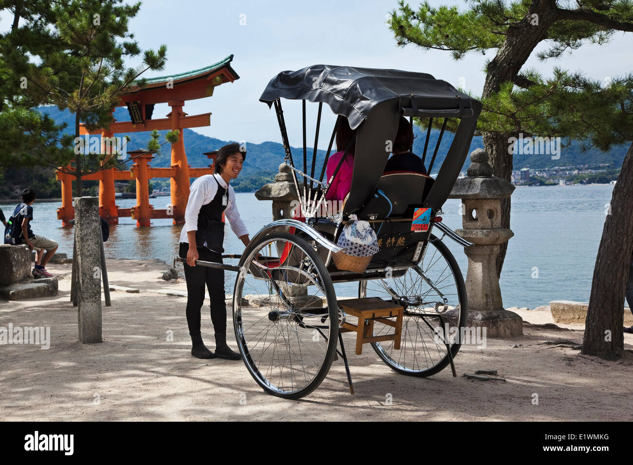 Smiling runner de parler avec les touristes assis dans son pousse-pousse sur l'île de Miyajima au Japon. Dans l'arrière-plan est la porte ou torii Banque D'Images