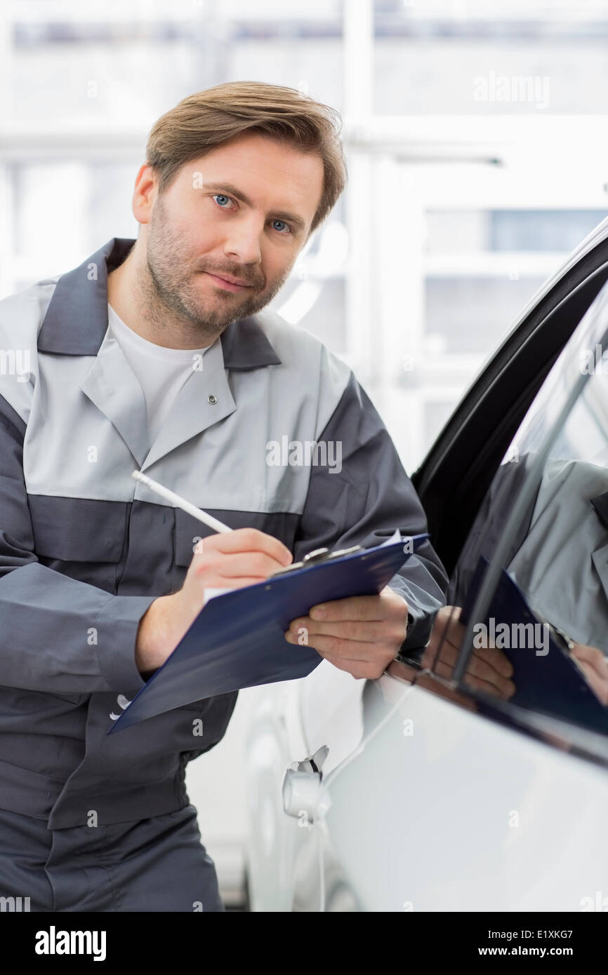 Portrait of smiling mechanic holding clipboard tout en s'appuyant sur la fenêtre de voiture en atelier Banque D'Images