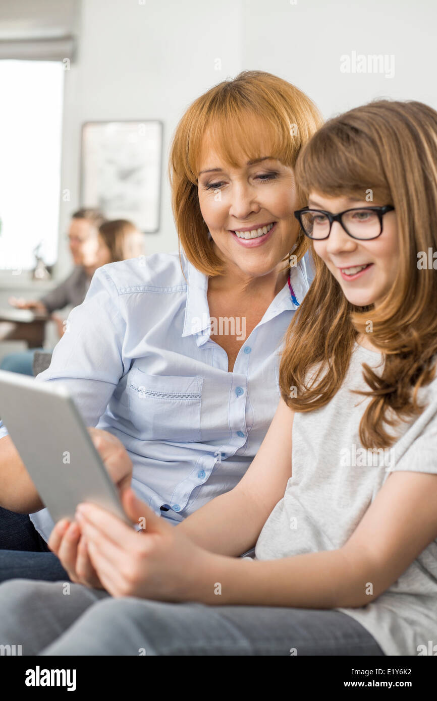 Happy mother and daughter using tablet PC avec la famille assis en arrière-plan à la maison Banque D'Images