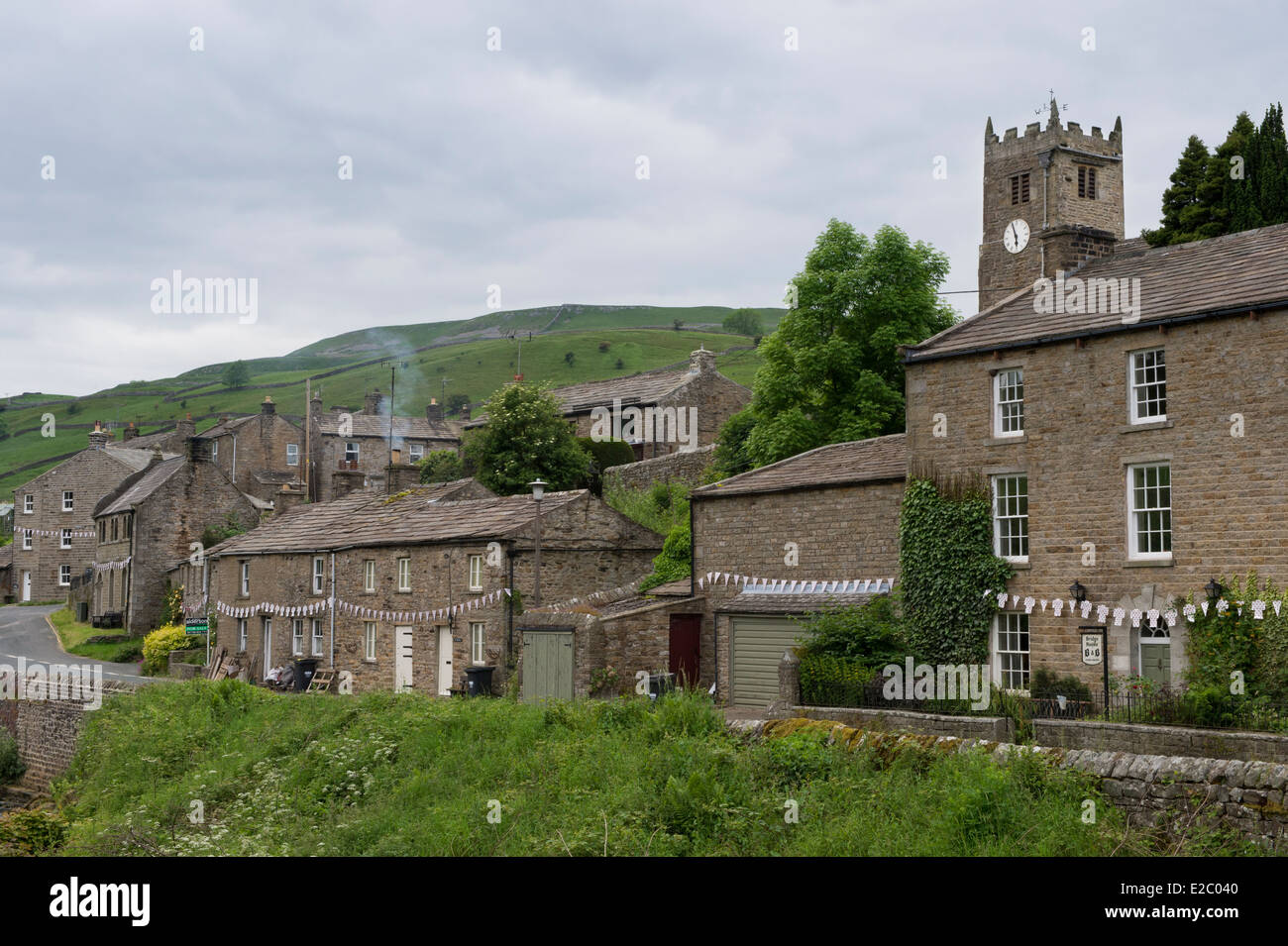 Chalets traditionnels en pierre bordant la route, tour de l'église et les collines au-delà - Muker, petit village rural de Swaledale, Yorkshire, Angleterre, Royaume-Uni. Banque D'Images