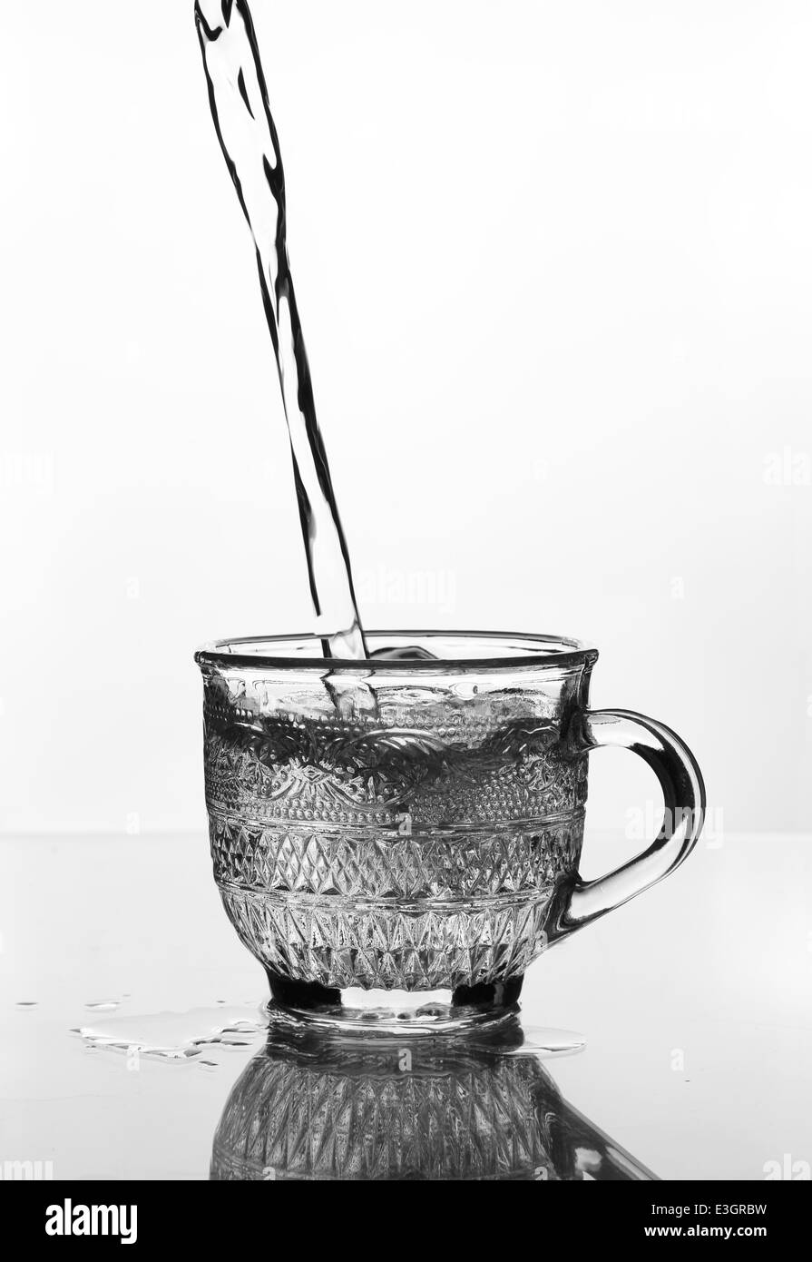 Verre tasse avec de l'eau verser sur une table en verre (échelle de gris) Banque D'Images