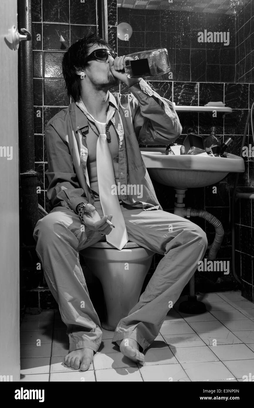 L'homme ivre est assis dans une toilette boire du whisky et fumer, image en niveaux de gris Banque D'Images