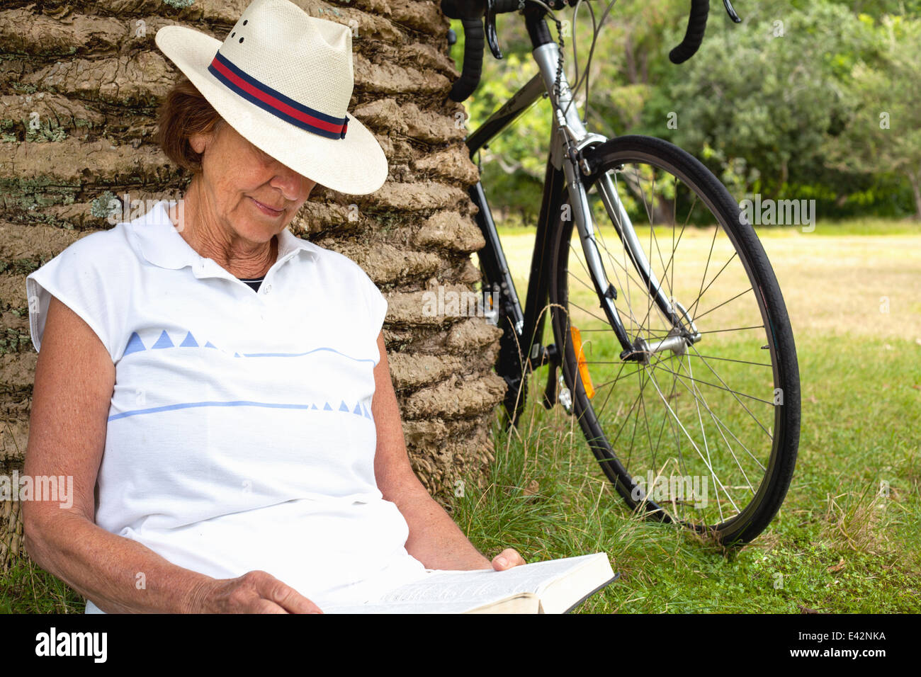 Senior woman leaning against palm tree reading in park Banque D'Images