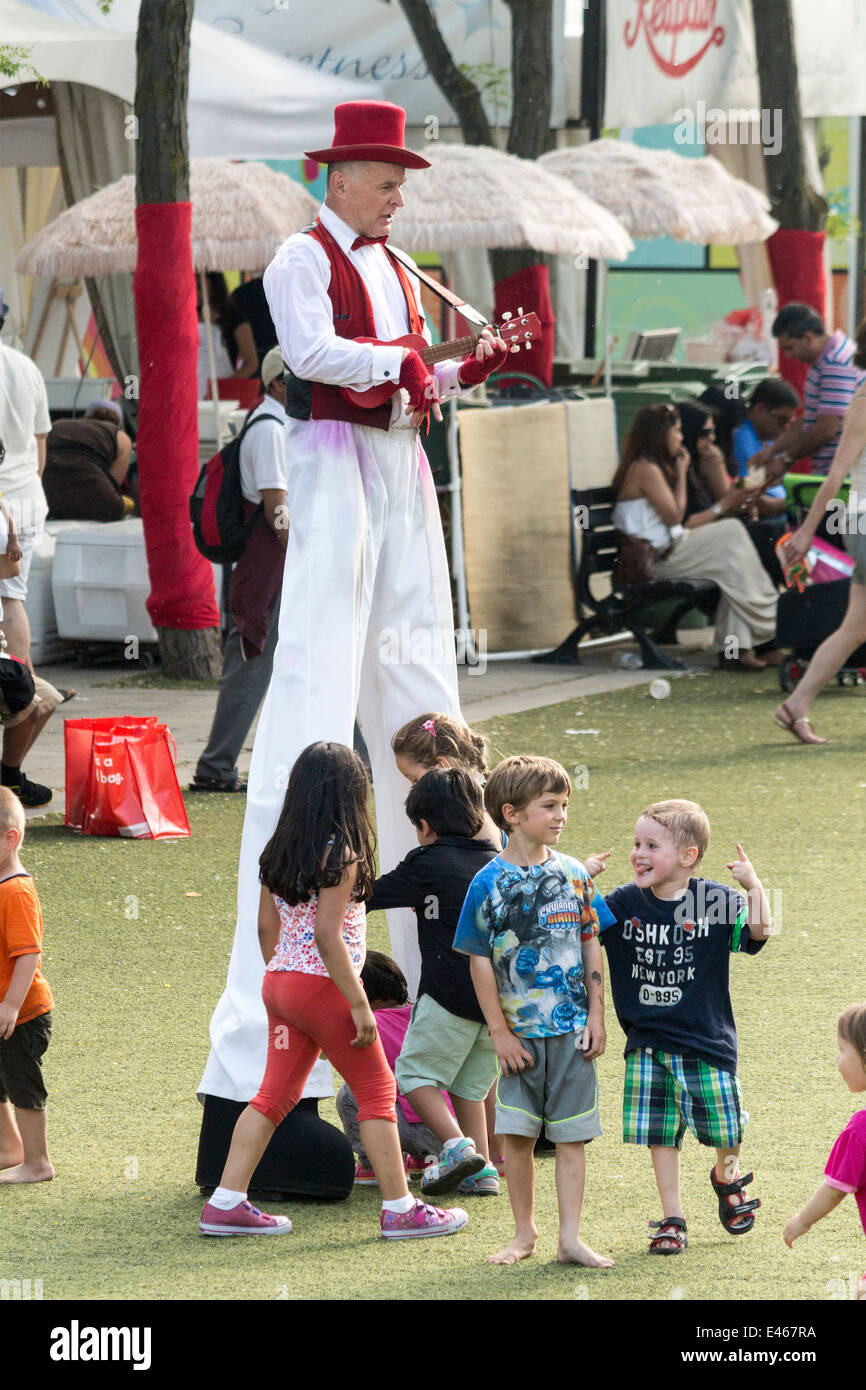 L'homme sur des échasses habillé en rouge et blanc à l'ukulélé alors que les enfants courir entre les jambes Banque D'Images