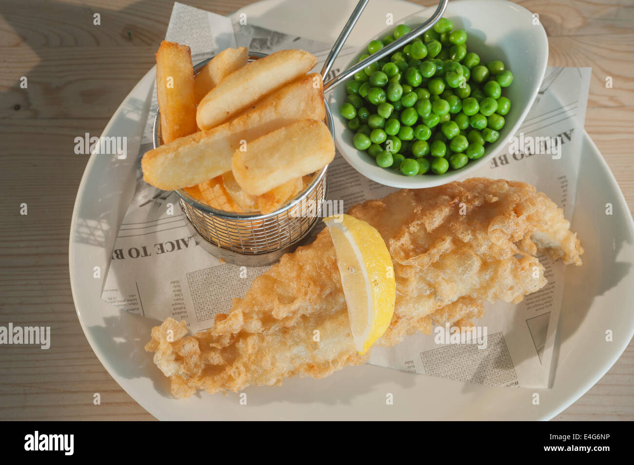 Fish and chips (aiglefin en pâte frits) avec des petits pois et un segment de citron sur une plaque blanche sur une table en bois Banque D'Images