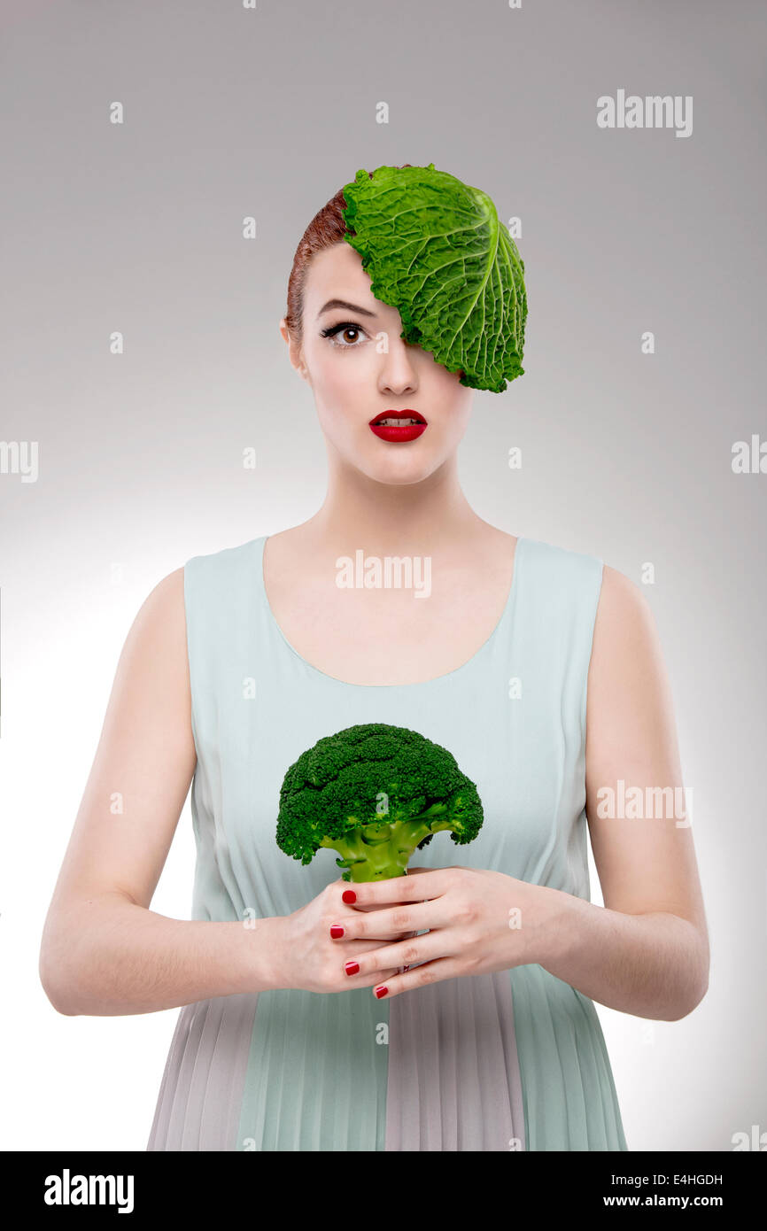 Portrait d'une femme végétalienne illustrant un concept avec un chou sur la tête et tenant un broccoli Banque D'Images