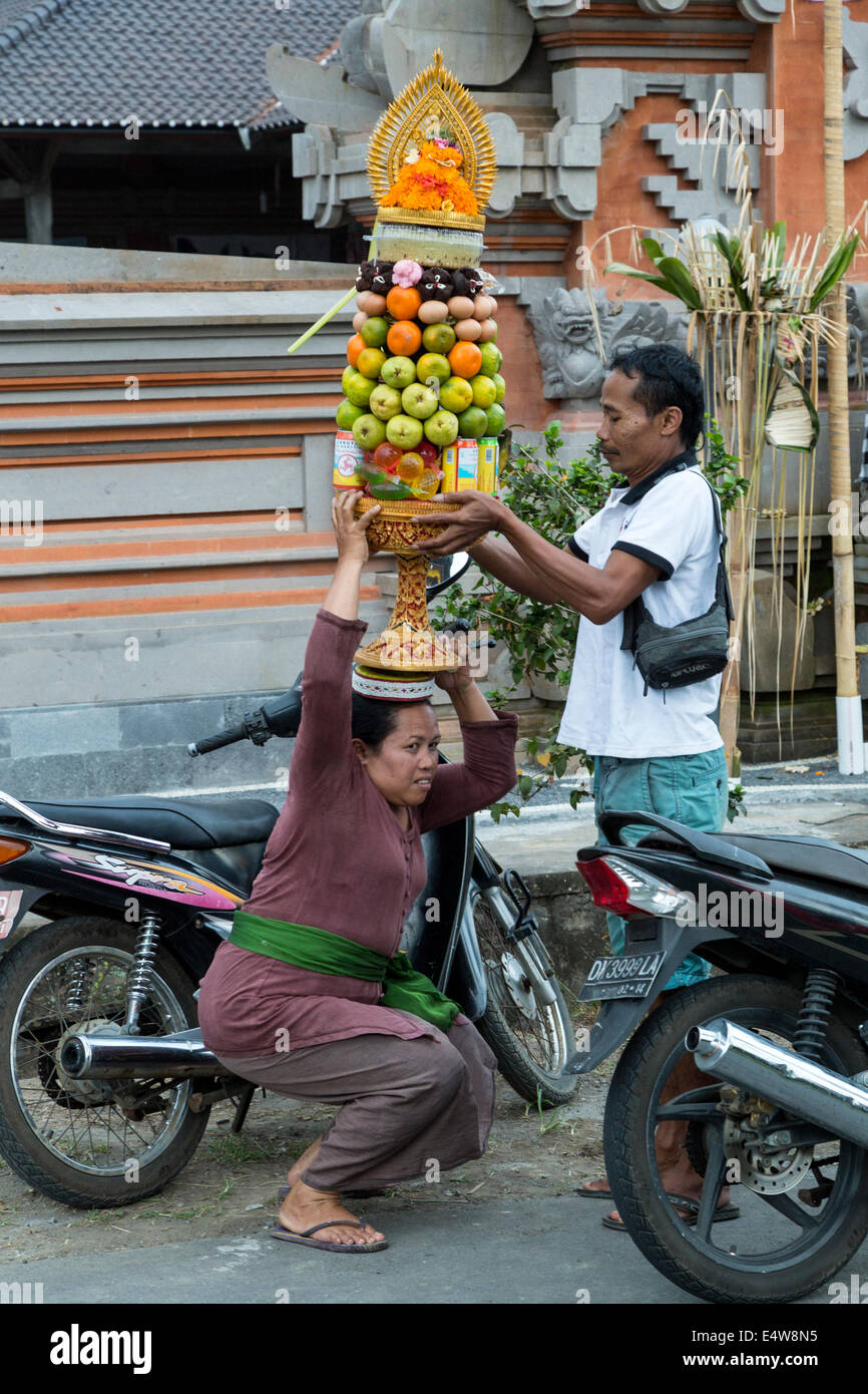 Bali, Indonésie. Homme Femme aide à équilibrer une offrande (Banten) sur sa tête avant d'entrer dans un temple. Banque D'Images
