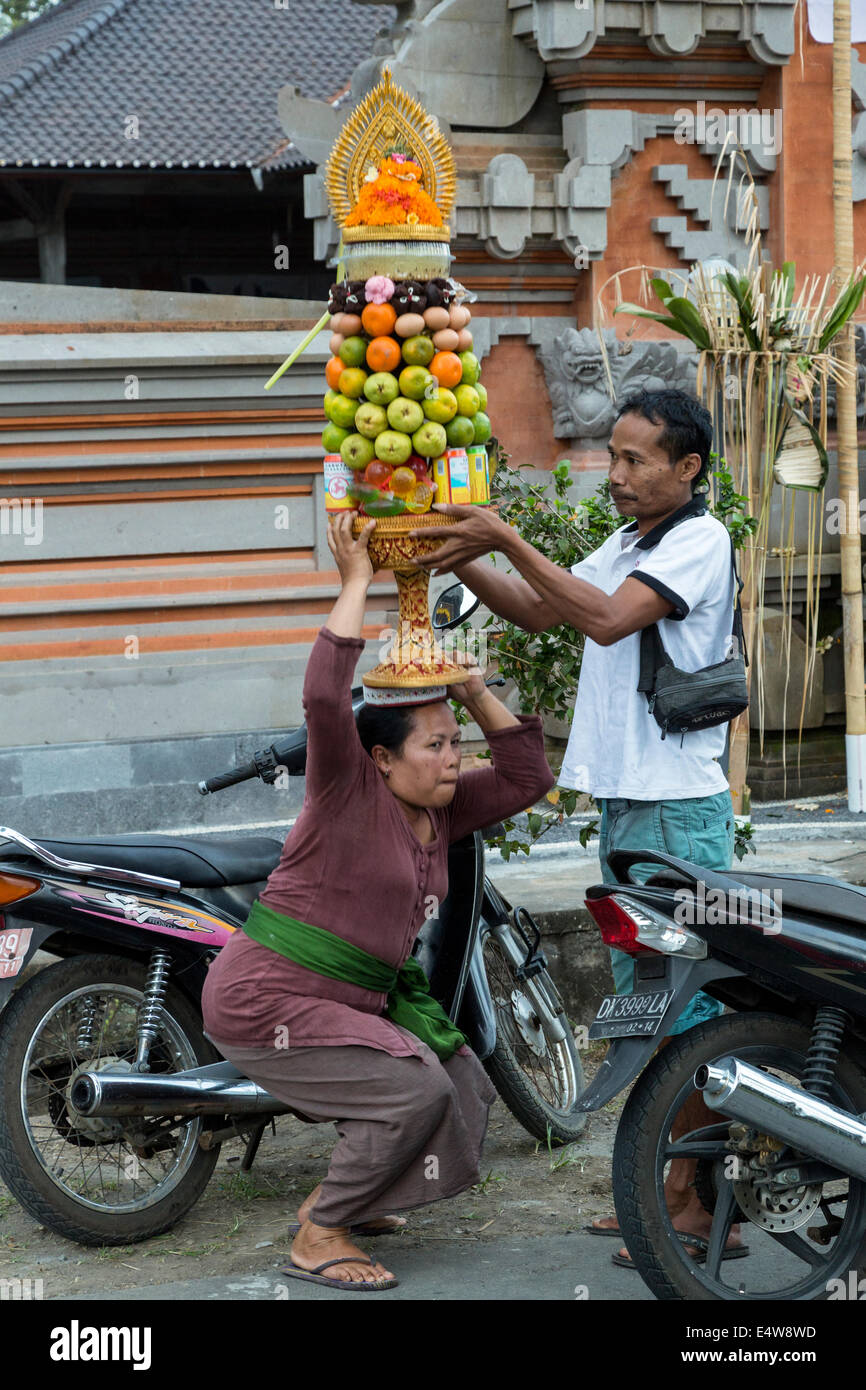 Bali, Indonésie. Homme Femme aide à équilibrer une offrande (Banten) sur sa tête avant d'entrer dans un temple. Banque D'Images