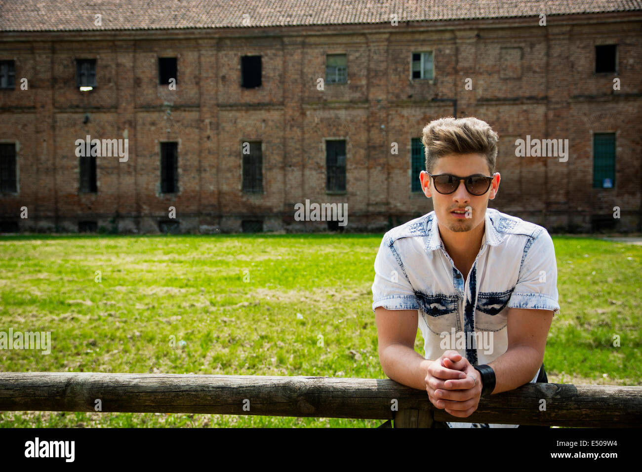 Beau jeune homme extérieur leaning on clôture en bois, ancienne maison de ferme ou derrière lui Banque D'Images