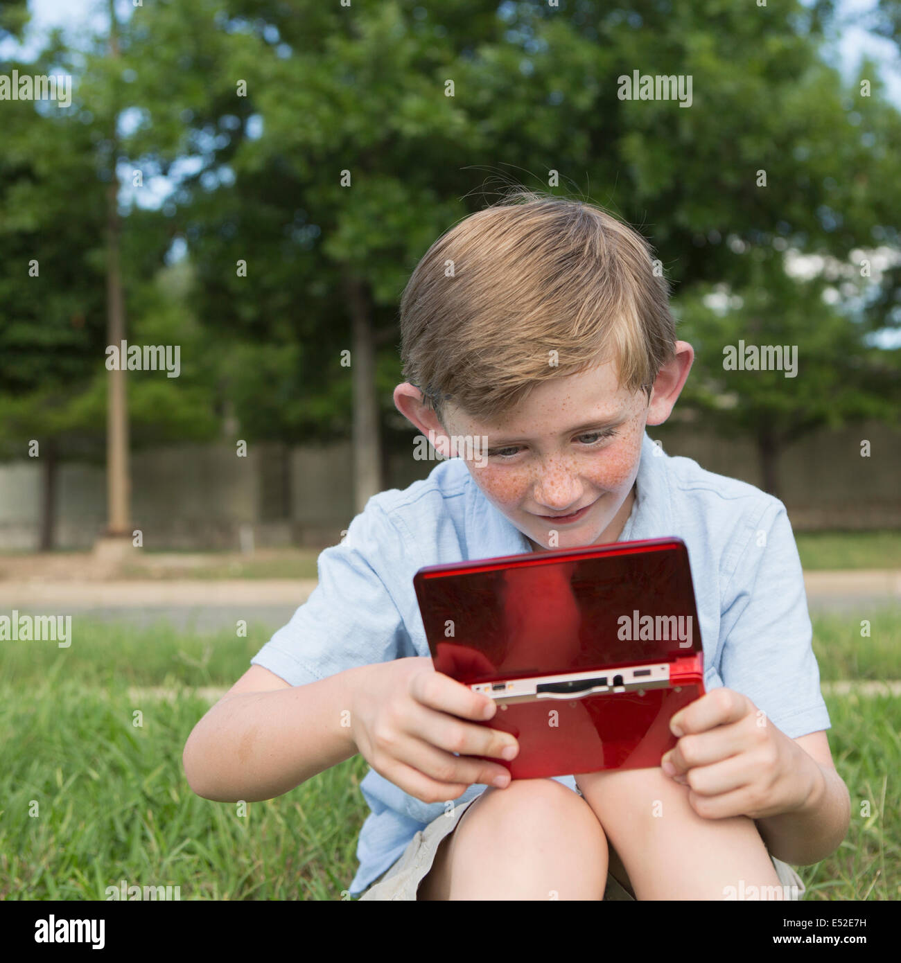 Un jeune garçon assis sur l'herbe de la lecture d'un jeu électronique de poche. Banque D'Images