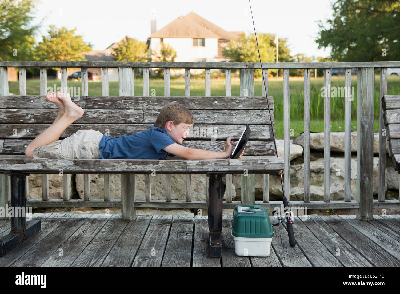 Un jeune garçon allongé sur un banc à l'extérieur à l'aide d'une tablette numérique. du matériel de pêche. Banque D'Images