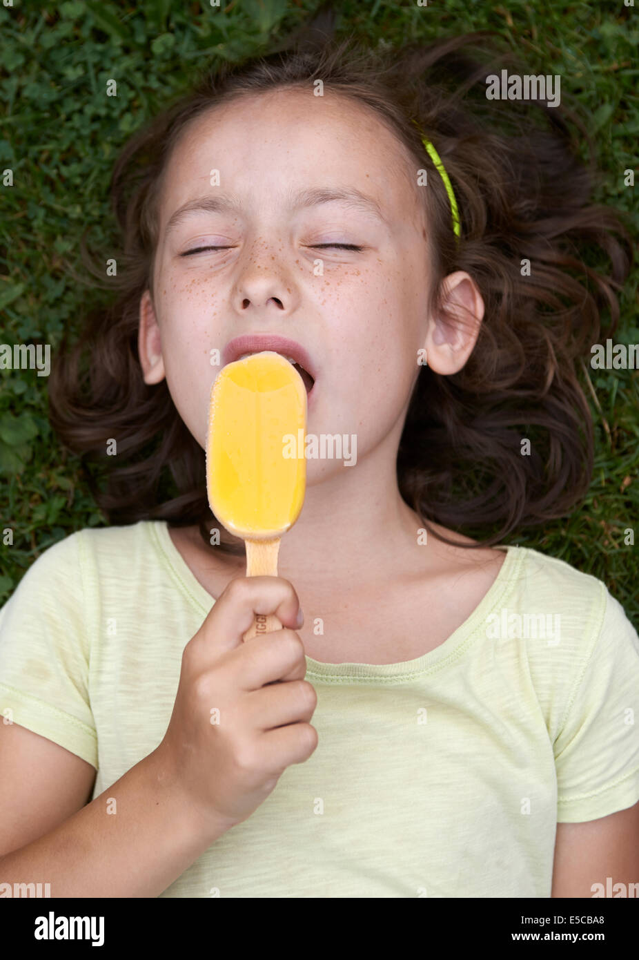 Enfant fille brune foncé léchant une glace, portrait, fond d'herbe à gazon vert couché Banque D'Images