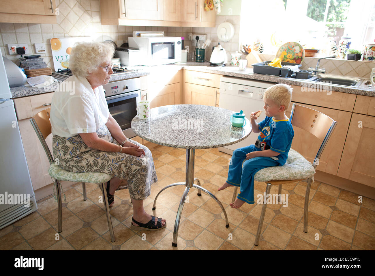Grand-mère assise à la table avec son petit-fils (de 3 ans) alors qu'il boit une tasse de lait et mange un biscuit, Royaume-Uni Banque D'Images