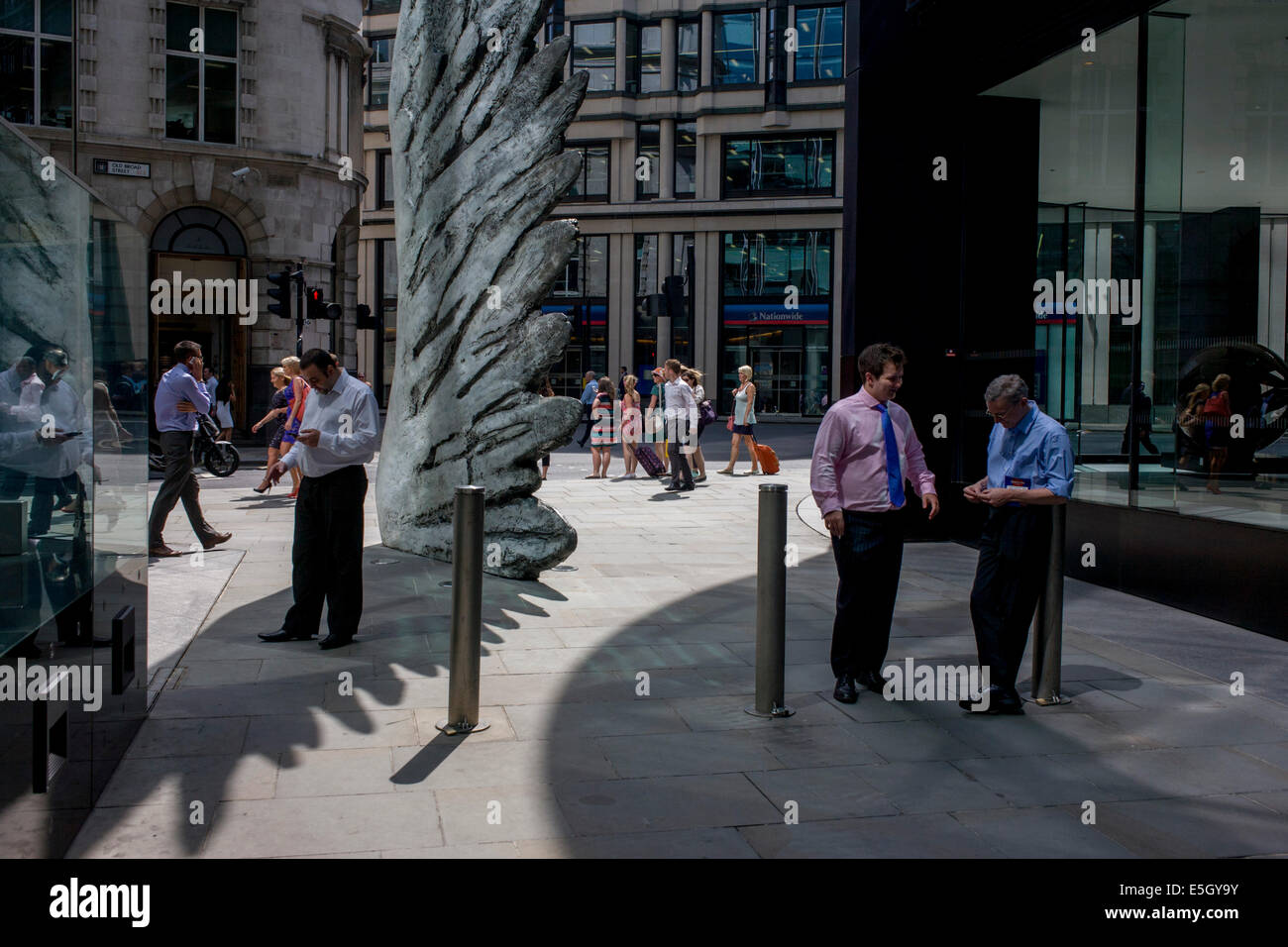 Les travailleurs de la ville de fumer des cigarettes par le géant de l'oeuvre une aile de bronze lors d'un midi de printemps dans le quartier financier de Londres. Comme la lumière se reflète sur les immeubles de bureaux à proximité, la foule à l'heure du déjeuner devant ce géant des illustrations sur leur façon de réunions et sandwicheries. Les dix mètres de haut est la sculpture en bronze par le président de la Royal Academy of Arts, Christopher Le Brun, commandé par Hammerson en 2009. Elle est appelée 'La Ville' de l'aile et a été jeté par Morris Singer, fondateurs de l'art réputé pour être la plus ancienne fonderie d'art dans le monde. Banque D'Images