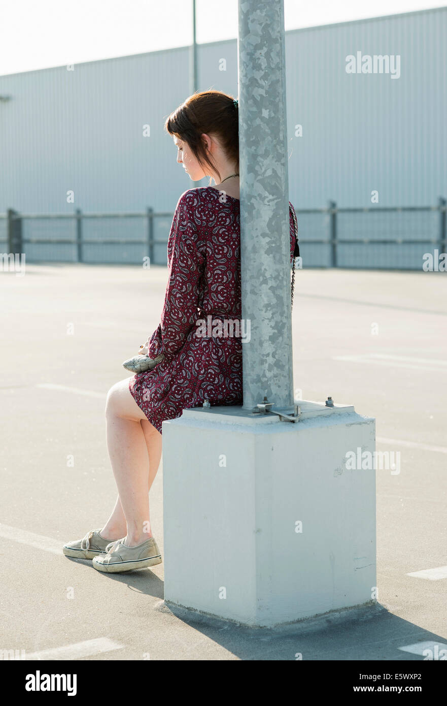 Jeune femme assise appuyé contre un lampadaire dans parking vide Banque D'Images